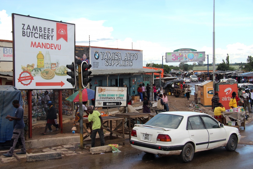 Ein Foto von einer Straßenszene in Sambia: Ein Auto, Passanten und einige Werbetafeln.