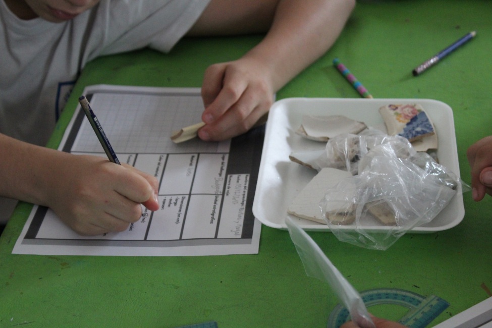 Das Foto zeigt eine Kinderhand, die in der linken Hand eine Tonscherbe hält und mit der rechten eine Notiz auf einem Papier verfasst.