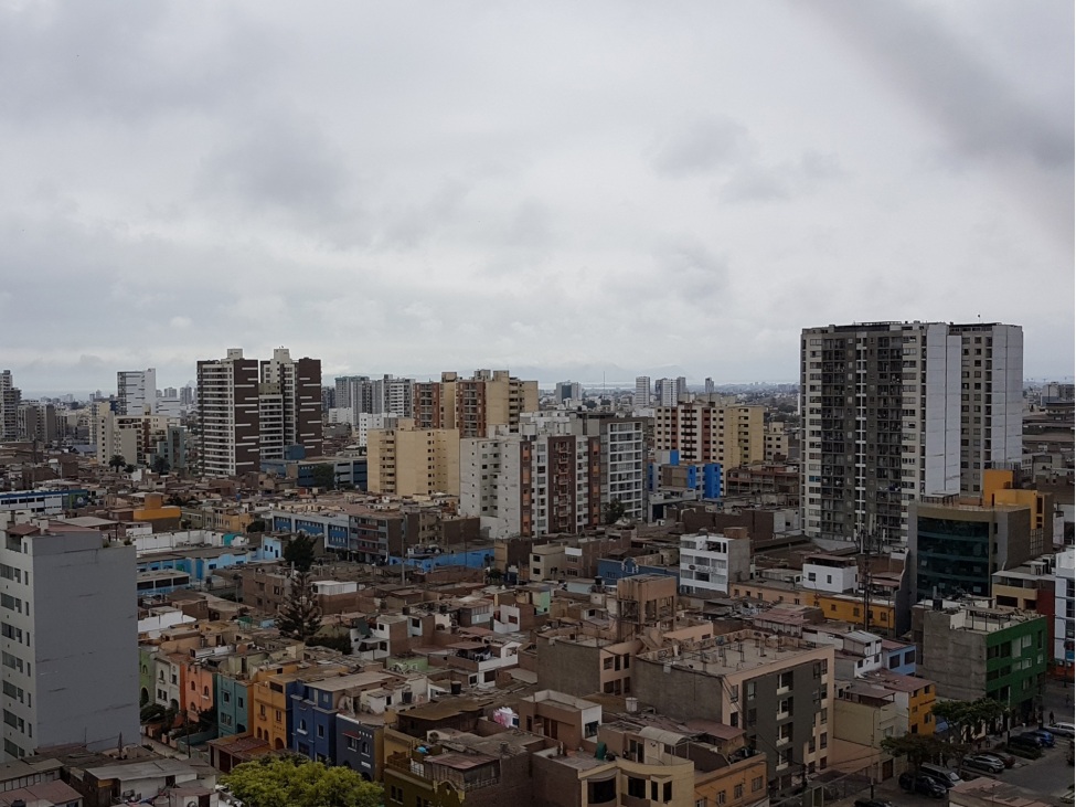 Eine Fotoaufnahme auf eine Stadt. Im Vordergrund sind farbige 3-5-geschossige Gebäude zu erkennen. Im Bildzentrum stehen vor allem Hochhäuser. Der Himmel ist bewölkt.