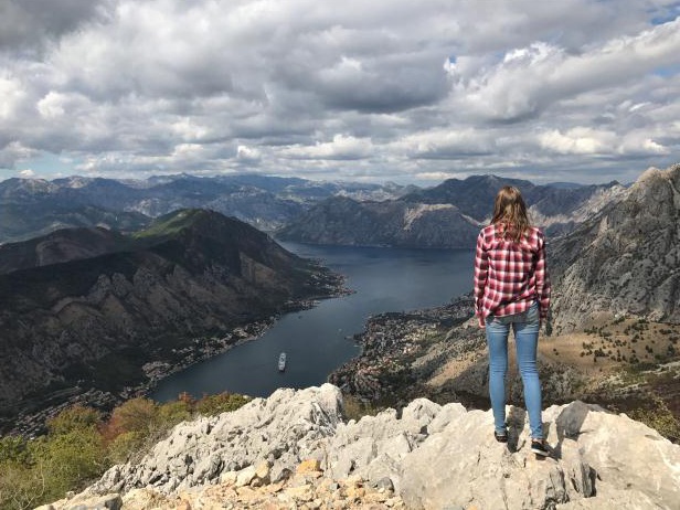 Ein Foto mit einer jungen Person von hinten, die an einem Abhang steht. Im Hintergrund ist die Berglandschaft und im Tal ein Gewässer zu erkennen.