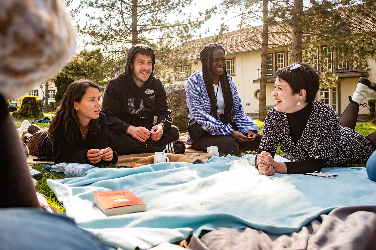 Eine Gruppe von fünf jungen Erwachsenen sitzt entspannt auf einer Decke im Freien und genießt ein Picknick. Im Vordergrund sind eine Flasche Wasser und ein Buch zu sehen. Die Gruppe unterhält sich angeregt und lacht. Im Hintergrund sind Bäume und ein Gebäude zu sehen. Die Atmosphäre wirkt warm und freundlich.