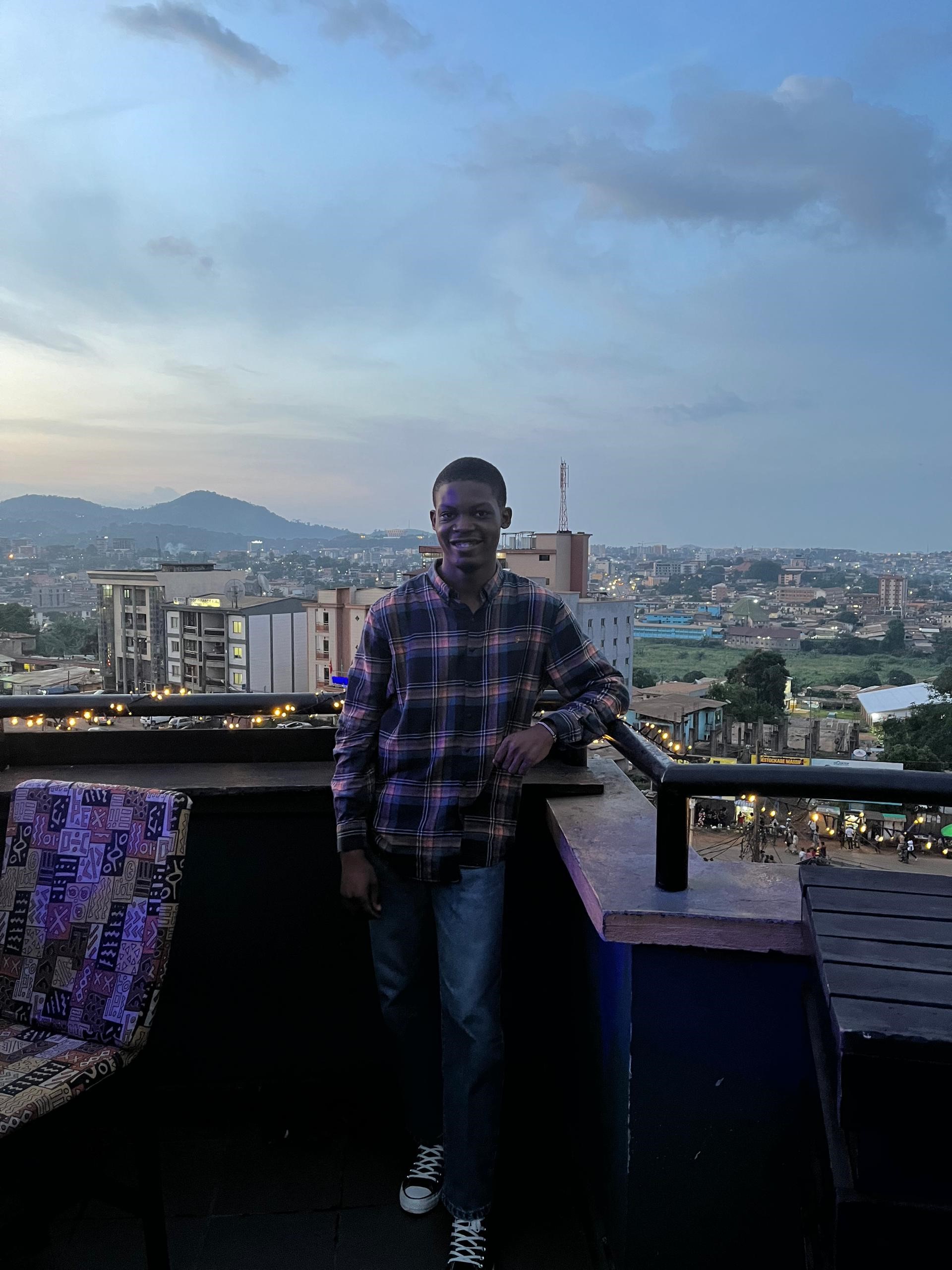 Auf dem Foto lehnt sich Alex in einem karierten Hemd und Jeans an einem Balkongeländer mit Stadtansicht und Hügeln im Hintergrund bei Sonnenuntergang. Der Balkon ist mit Lichterketten geschmückt und ein bunter Stuhl steht daneben.