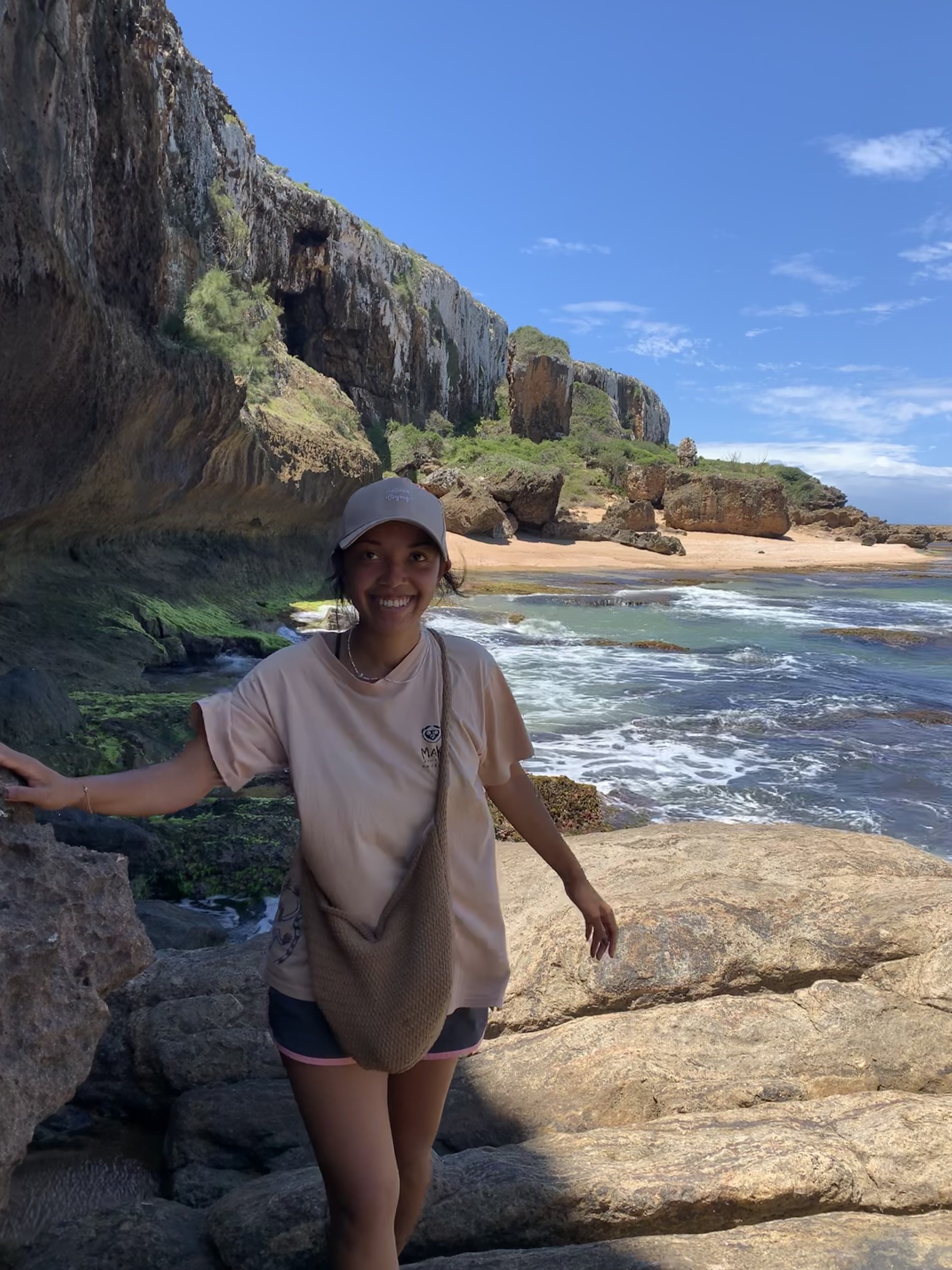 Anne steht in einem hellen T-Shirt und einer Kappe auf felsigem Gelände an einem Küstestreifen. Sie hält sich an einem Felsen fest, während im Hintergrund eine malerische Klippenlandschaft und das Meer zu sehen sind. Der Himmel ist klar und blau.