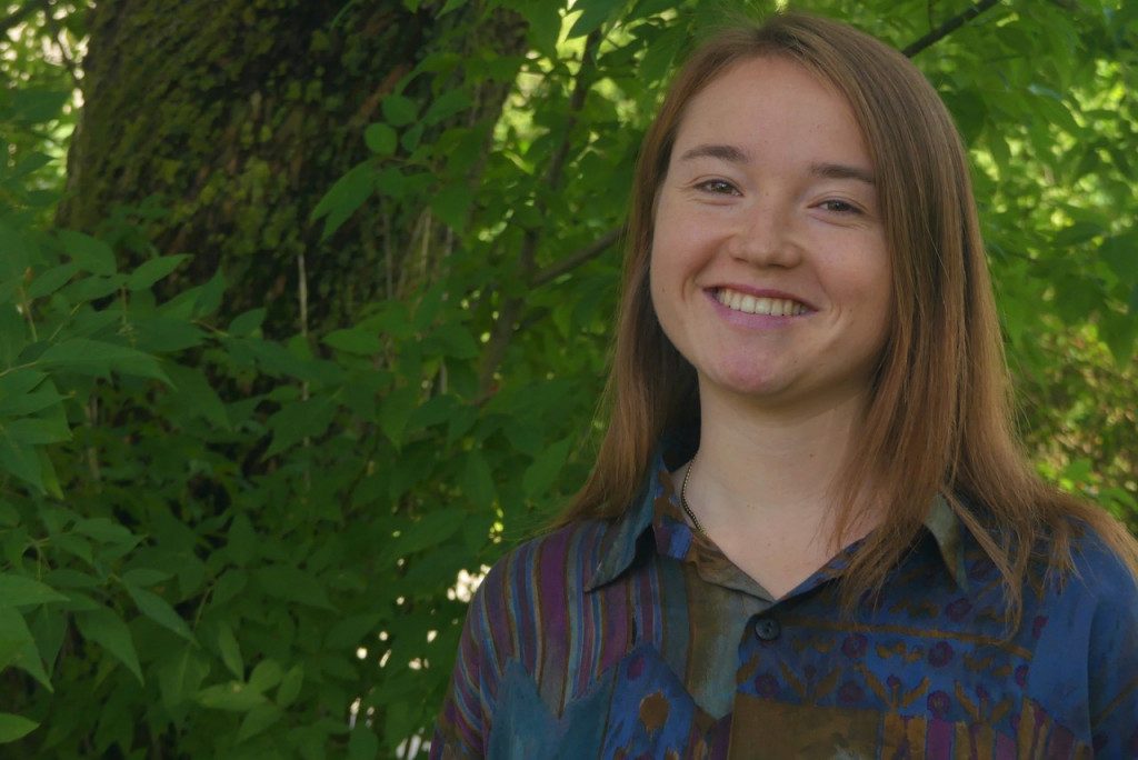 Das Bild zeigt Mara Urich. Sie hat lange, glatte, braune Haare und lächelt in die Kamera. Sie trägt ein bunt gemustertes Hemd. Im Hintergrund sind grüne Blätter und ein Baumstamm zu sehen, was auf eine Wald- oder Gartenumgebung hinweist.