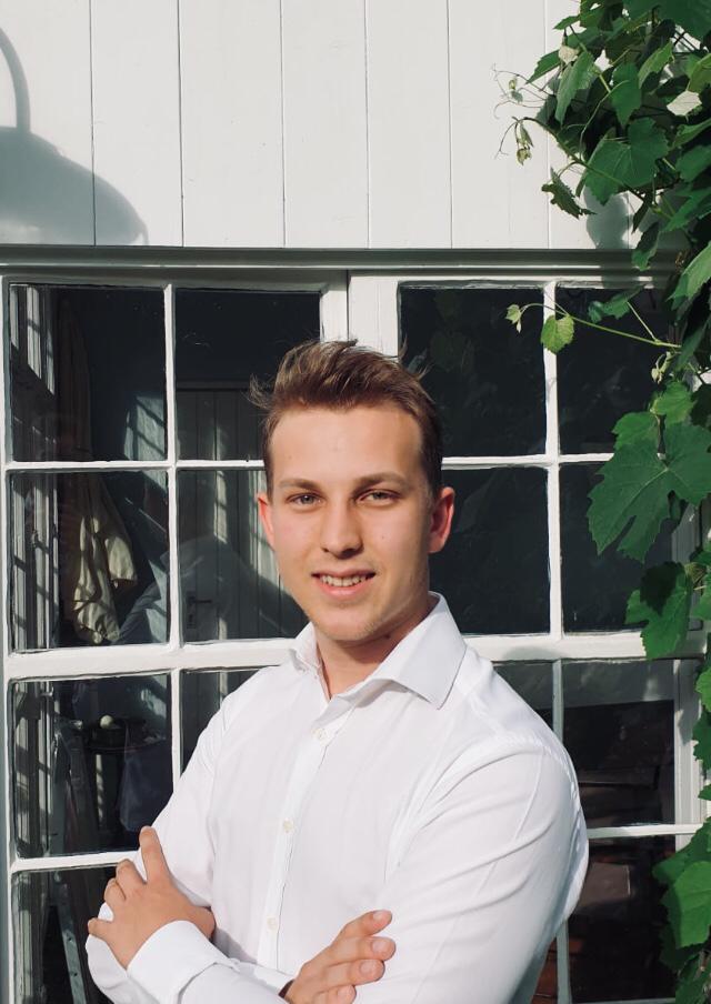 Das Bild zeigt Nelson Kück. Er hat kurze braune Haare und trägt ein weißes Hemd. Er hat seine Arme verschränkt und lächelt in die Kamera. 