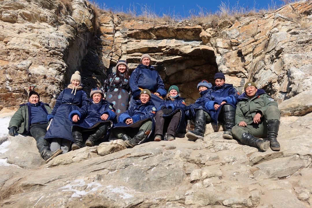 Das Bild zeigt eine Gruppe von Menschen, die auf Felsen vor einer kleinen Höhle oder einem natürlichen Felsvorsprung sitzen. Die Personen sind warm gekleidet, tragen dicke Jacken, Mützen und Stiefel, was darauf hindeutet, dass es kalt ist. Alle lächeln in die Kamera und wirken entspannt und glücklich. Der Hintergrund zeigt eine felsige Landschaft mit etwas Gras an den oberen Kanten der Felsen und einem strahlend blauen Himmel. Die Szene strahlt eine ruhige, freundliche Atmosphäre in einer rauen, aber schöne