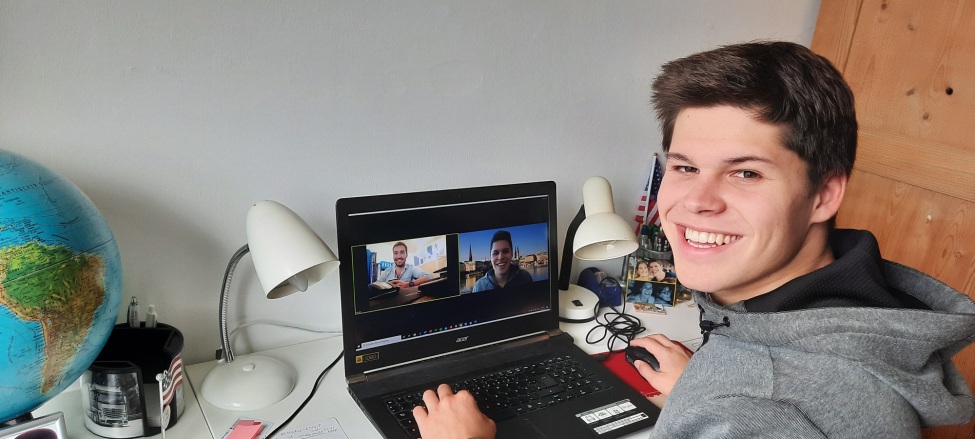 Das Bild zeigt Paulo Glowacki, der an einem Schreibtisch sitzt und in die Kamera lächelt, während er an einem Videoanruf auf seinem Laptop teilnimmt. Auf dem Bildschirm des Laptops sind zwei weitere Personen zu sehen, die ebenfalls in den Videoanruf involviert sind. 