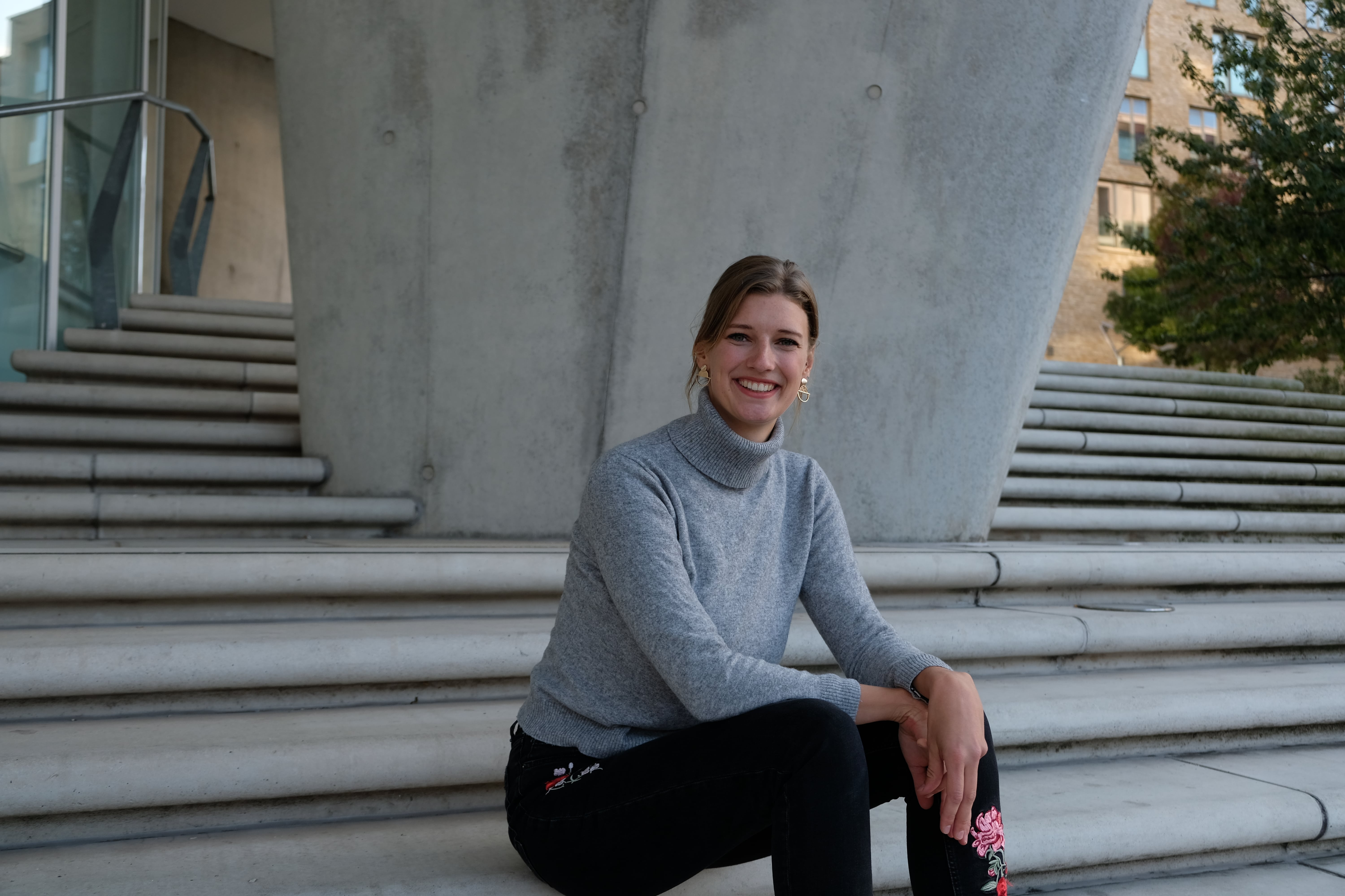 Das Bild zeigt die Alumna Bettina Gschwend. Sie trägt ihre Haare zusammengebunden und einen grauen Rollkragenpullover. Sie sitzt auf einer Betontreppe.