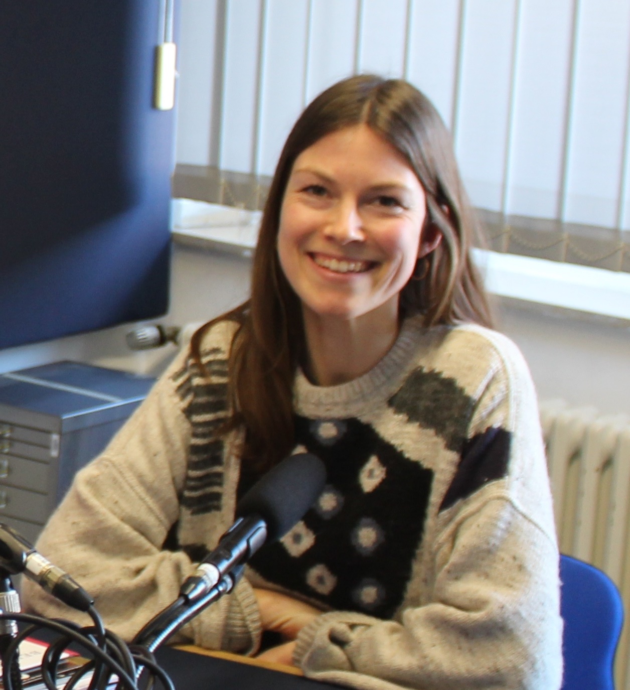 Das Bild zeigt Jessica Best, sie hat braune lange Haare und trägt einen schwarz-beige gemusterten Pullover.