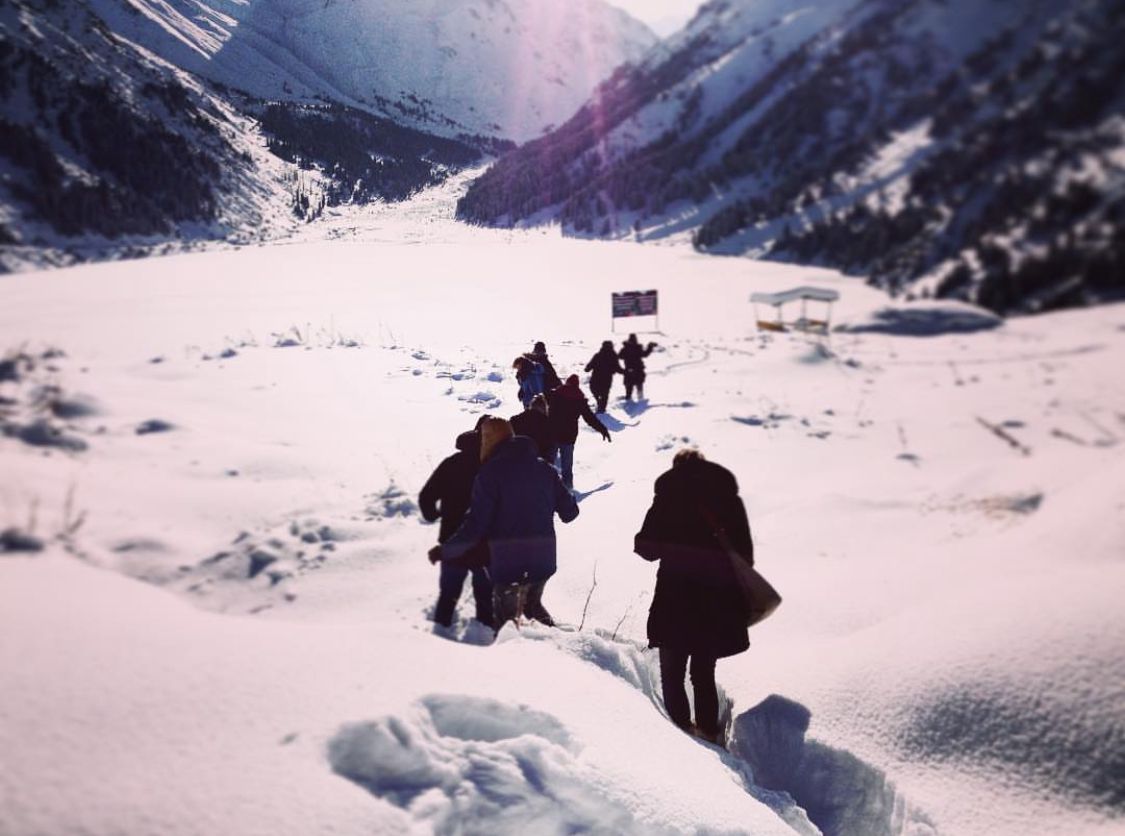 Das Bild zeigt eine Gruppe von Menschen, die in einer verschneiten Berglandschaft wandern. Sie folgen einem schmalen Pfad durch tiefen Schnee, der durch die unberührte, weiße Schneedecke führt. Die Berge um sie herum sind hoch und imposant, mit steilen Hängen und ebenfalls mit Schnee bedeckt. Die Sonne scheint hell, wodurch die Schneelandschaft noch intensiver leuchtet, und lange Schatten auf den Schnee wirft. Die Gruppe ist warm gekleidet, was auf die kalten Temperaturen hinweist.