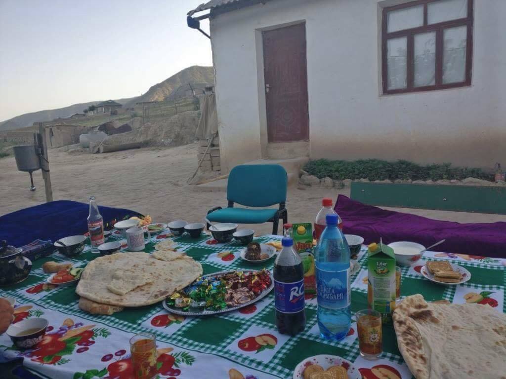 Das Bild zeigt einen Tisch im Freien, der mit einer grünen, karierten Tischdecke gedeckt ist. Auf dem Tisch liegen verschiedene Speisen und Getränke, darunter Fladenbrot, Süßigkeiten, Obst, Wasserflaschen, eine Flasche Cola, Teegläser und weitere traditionelle Speisen. Die Umgebung wirkt ländlich und einfach, mit einem kleinen weißen Haus im Hintergrund. 