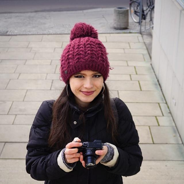 Das Bild zeigt Clara Hoheisel, die eine Kamera in den Händen hält und in die Kamera lächelt. Sie trägt eine warme, gestrickte Mütze in einem kräftigen Burgunderrot mit einem Pompon oben drauf sowie eine schwarze Winterjacke. Der Hintergrund besteht aus einer gepflasterten Straße oder einem Gehweg, der leicht verschwommen ist, was den Fokus auf Clara legt. 