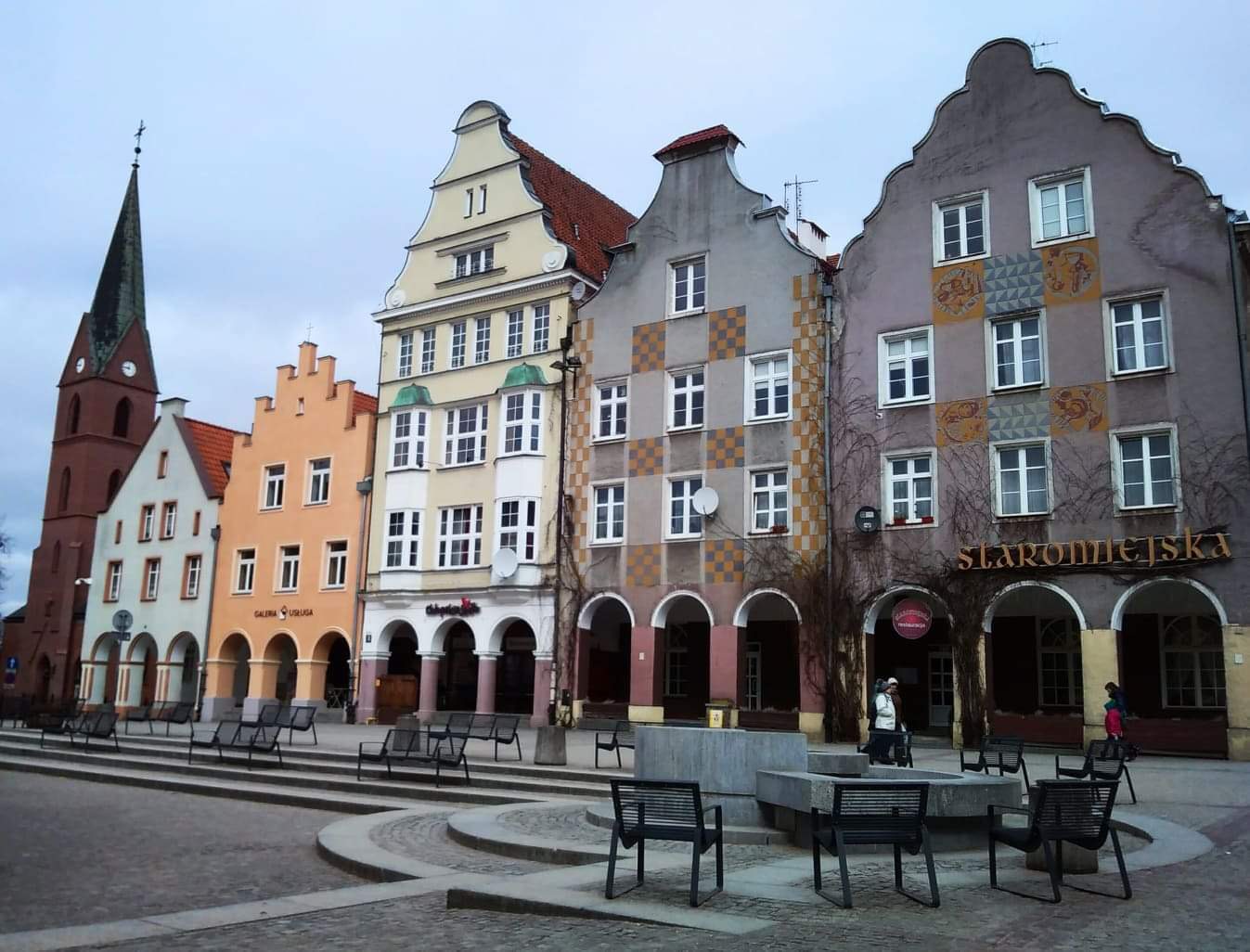 Das Bild zeigt eine malerische Stadtansicht mit einer Reihe von farbenfrohen, historischen Gebäuden, die in einem typisch europäischen Stil errichtet sind. Die Fassaden der Häuser sind in Pastellfarben wie Beige, Gelb, Orange und Lila gehalten, und sie sind mit dekorativen Elementen wie Wappen, Ornamenten und Giebeln verziert. 