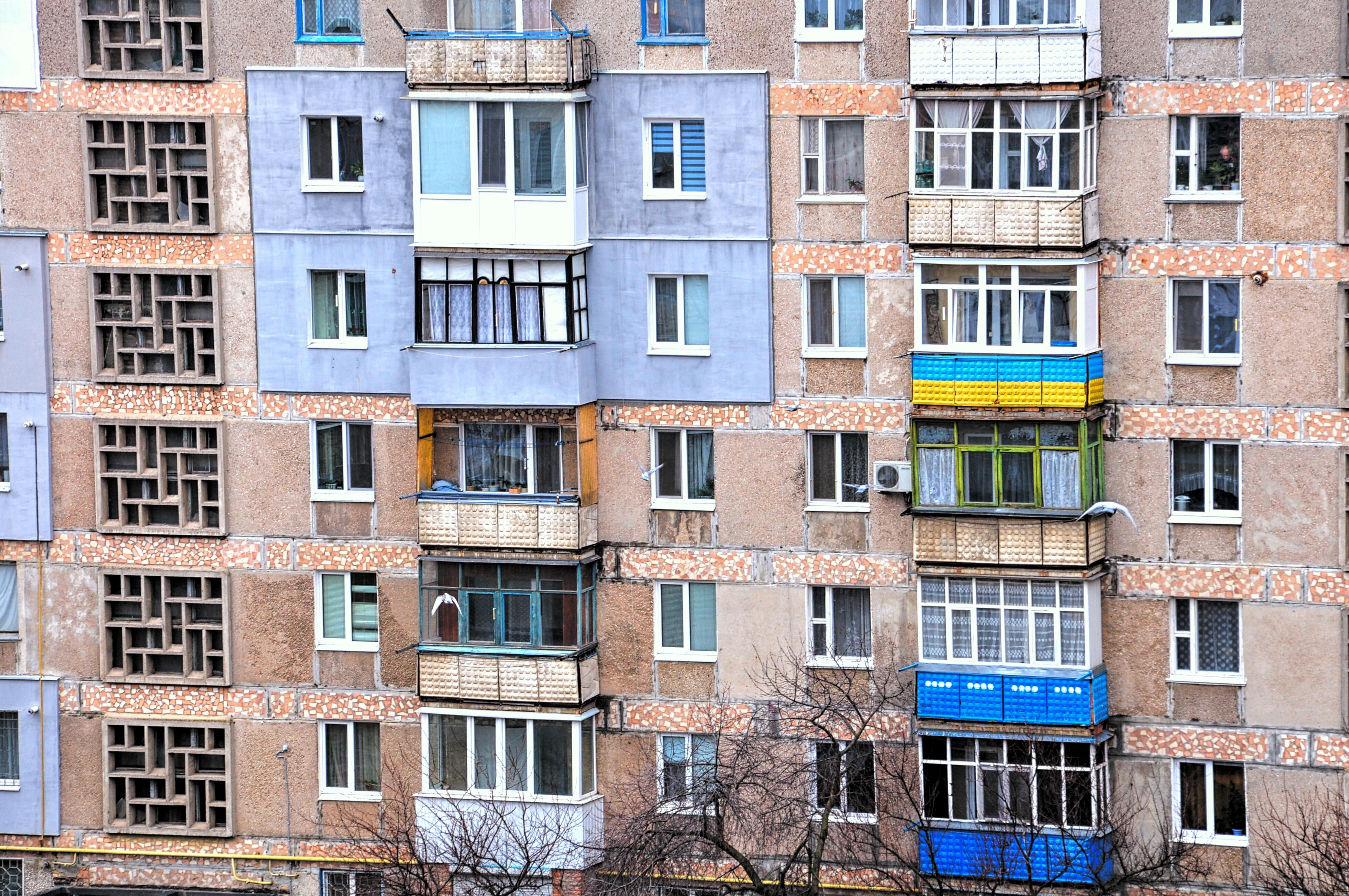 Das Bild zeigt die Fassade eines mehrstöckigen Wohngebäudes. Die Architektur ist typisch für ältere Plattenbauten, wie sie in vielen post-sowjetischen Ländern zu finden sind. Die Fassade ist in einem verwitterten Zustand, mit sichtbaren Abnutzungserscheinungen und partiellen Renovierungen. Die Balkone sind unterschiedlich gestaltet, einige sind verglast, andere offen.