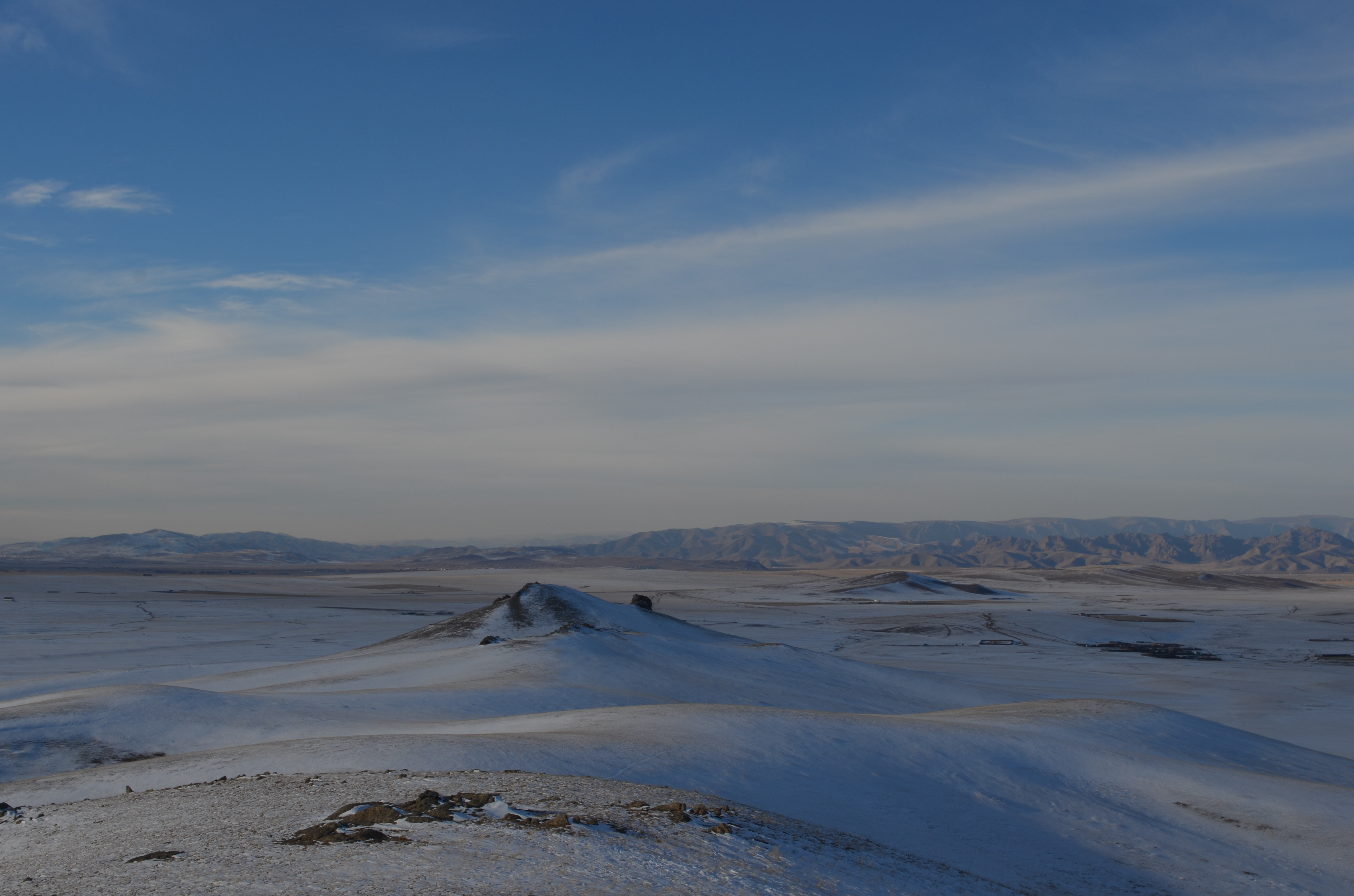 Das Bild zeigt eine weite, schneebedeckte Landschaft unter einem klaren, blauen Himmel. Im Vordergrund erstrecken sich sanfte Hügel, während am Horizont in der Ferne eine Gebirgskette zu sehen ist, die den weiten, unbewohnten Raum umrahmt. Die karge Schönheit der Landschaft vermittelt eine ruhige, aber auch kühle Atmosphäre.