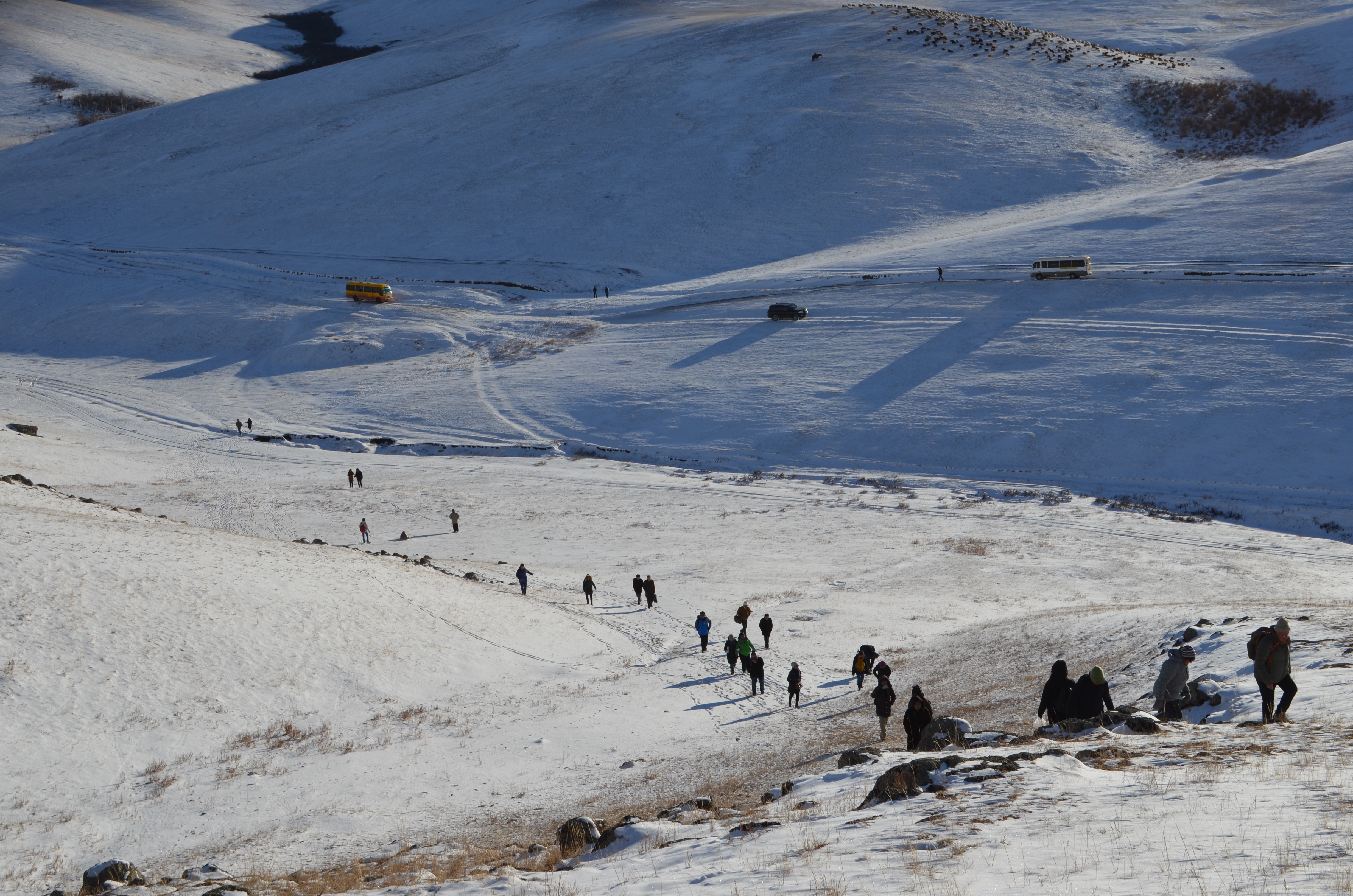 Das Bild zeigt eine Gruppe von Menschen, die einen schneebedeckten Hügel hinaufsteigen. Im Hintergrund sind zwei gelbe Fahrzeuge zu sehen, die auf einer Straße in der verschneiten Landschaft parken. Die Landschaft ist von sanften Hügeln geprägt, und das Licht der tiefstehenden Sonne wirft lange Schatten über den Schnee, was der Szene eine kühle, aber lebendige Atmosphäre verleiht.