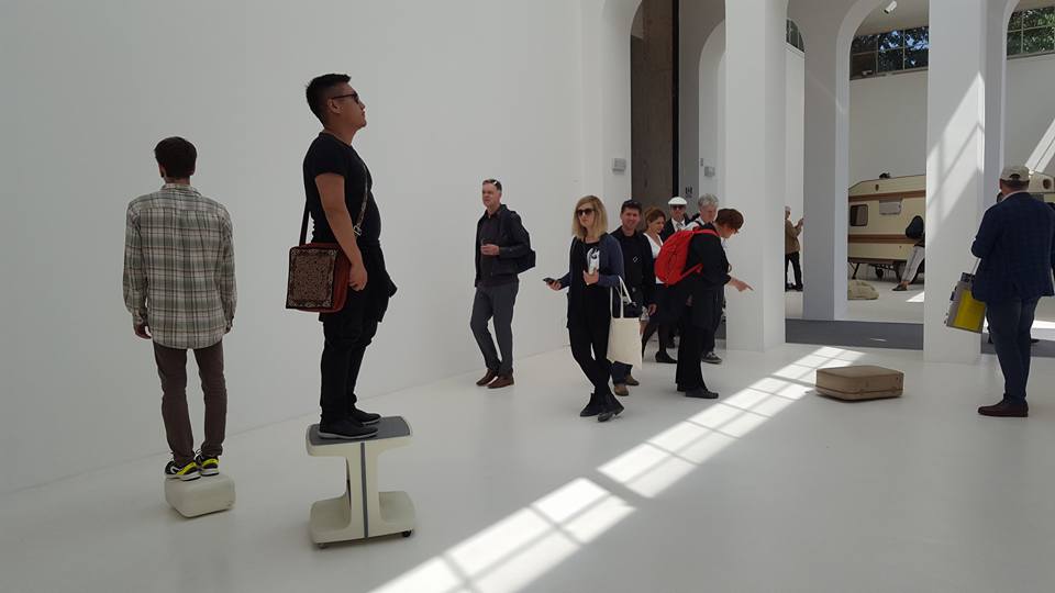 Das Bild zeigt eine Szene auf der Biennale in Venedig. Es ist eine moderne Ausstellungshalle mit weißen Wänden und einem Boden, auf dem Menschen stehen und gehen. Zwei Personen stehen auf kleinen Podesten und blicken in verschiedene Richtungen; eine Person ist dem Betrachter abgewandt, während die andere zur Seite schaut. Weitere Personen bewegen sich durch den Raum, einige betrachten Exponate, während andere miteinander sprechen oder ihre Mobiltelefone nutzen. 