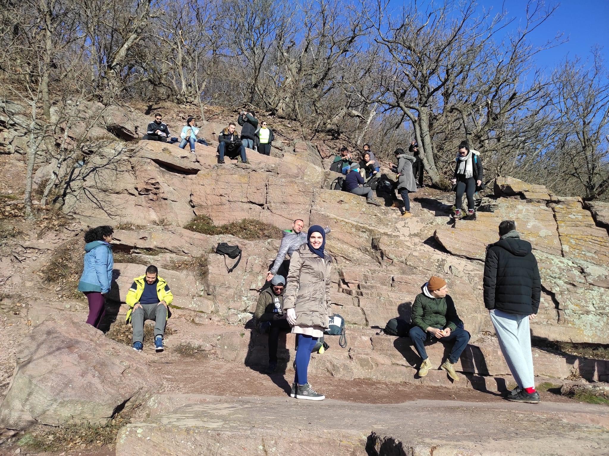 Das Bild zeigt eine Gruppe von Menschen, die auf einer felsigen Anhöhe sitzen und stehen, offensichtlich eine Pause während einer Wanderung machen. Die Felsen sind in verschiedenen Ebenen angeordnet, und die Menschen nutzen diese natürlichen "Sitzplätze", um sich auszuruhen, die Aussicht zu genießen oder miteinander zu sprechen. Einige der Personen tragen Winterkleidung, was auf kühles Wetter hinweist.