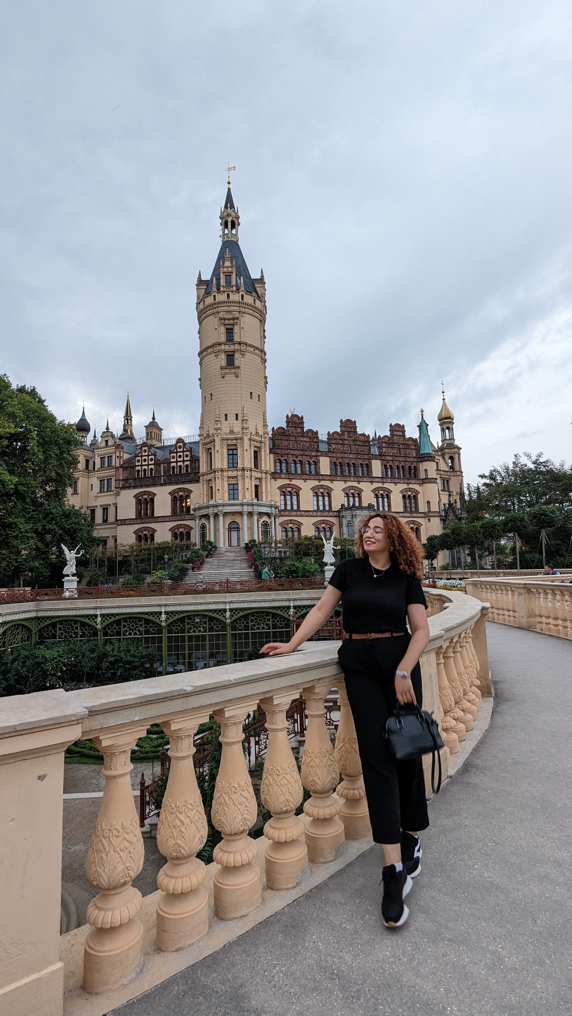 Das Bild zeigt Oumaima Nouali, die auf einer Brücke vor einem beeindruckenden Schloss posiert. Das Schloss im Hintergrund hat eine prächtige Architektur mit hohen Türmen, verzierten Fassaden und einer Mischung aus gotischen und Renaissance-Stilelementen. 