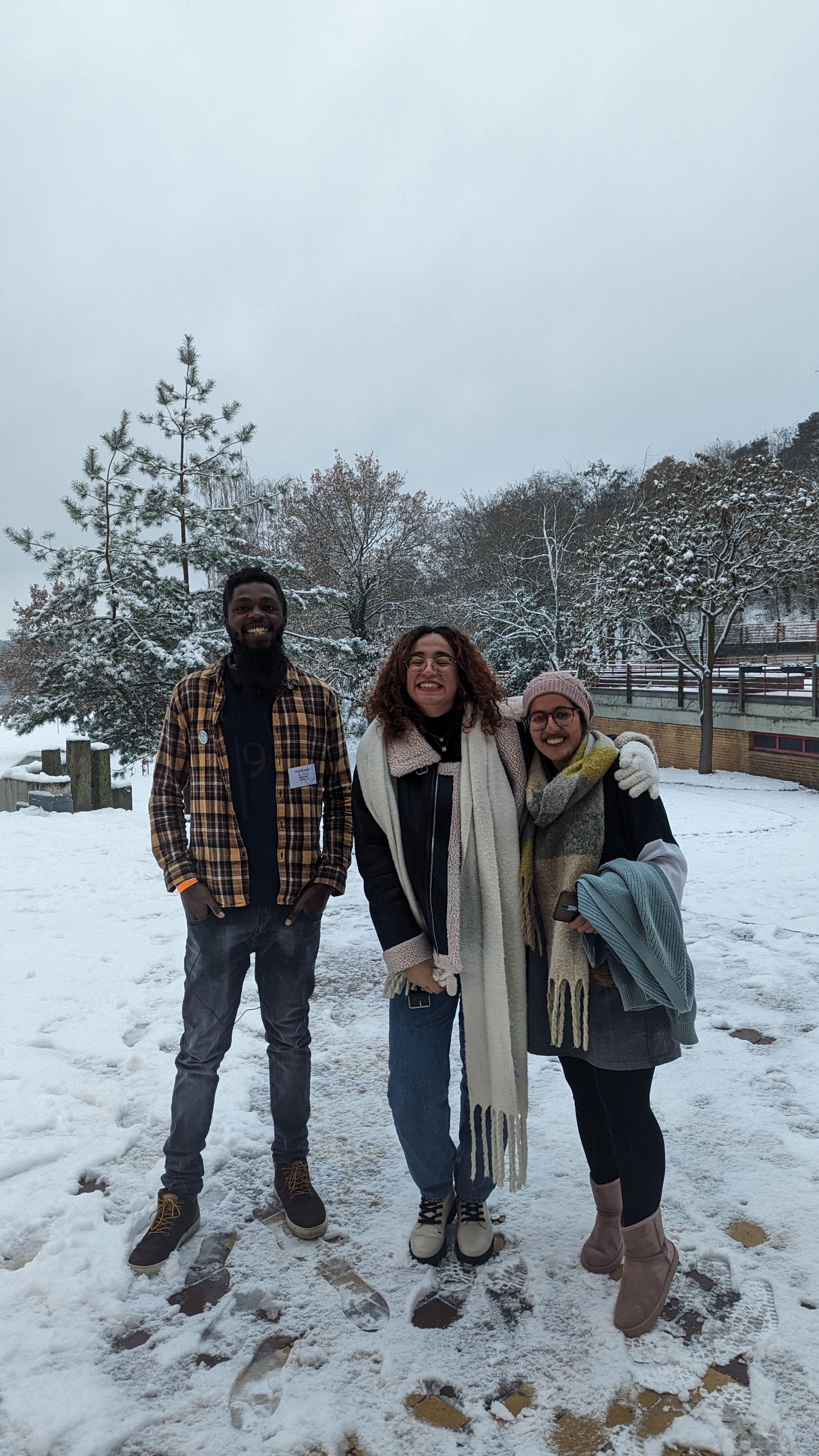 Das Bild zeigt drei Personen, die in einer verschneiten Umgebung stehen und fröhlich in die Kamera lächeln. In der Mitte steht Oumaima Nouali, eingehüllt in einen langen Schal und eine dicke Winterjacke. Links von ihr steht ein Mann in einer karierten Jacke und Jeans, und rechts eine Frau, die ebenfalls warm angezogen ist, mit einem Schal und einer Mütze. 