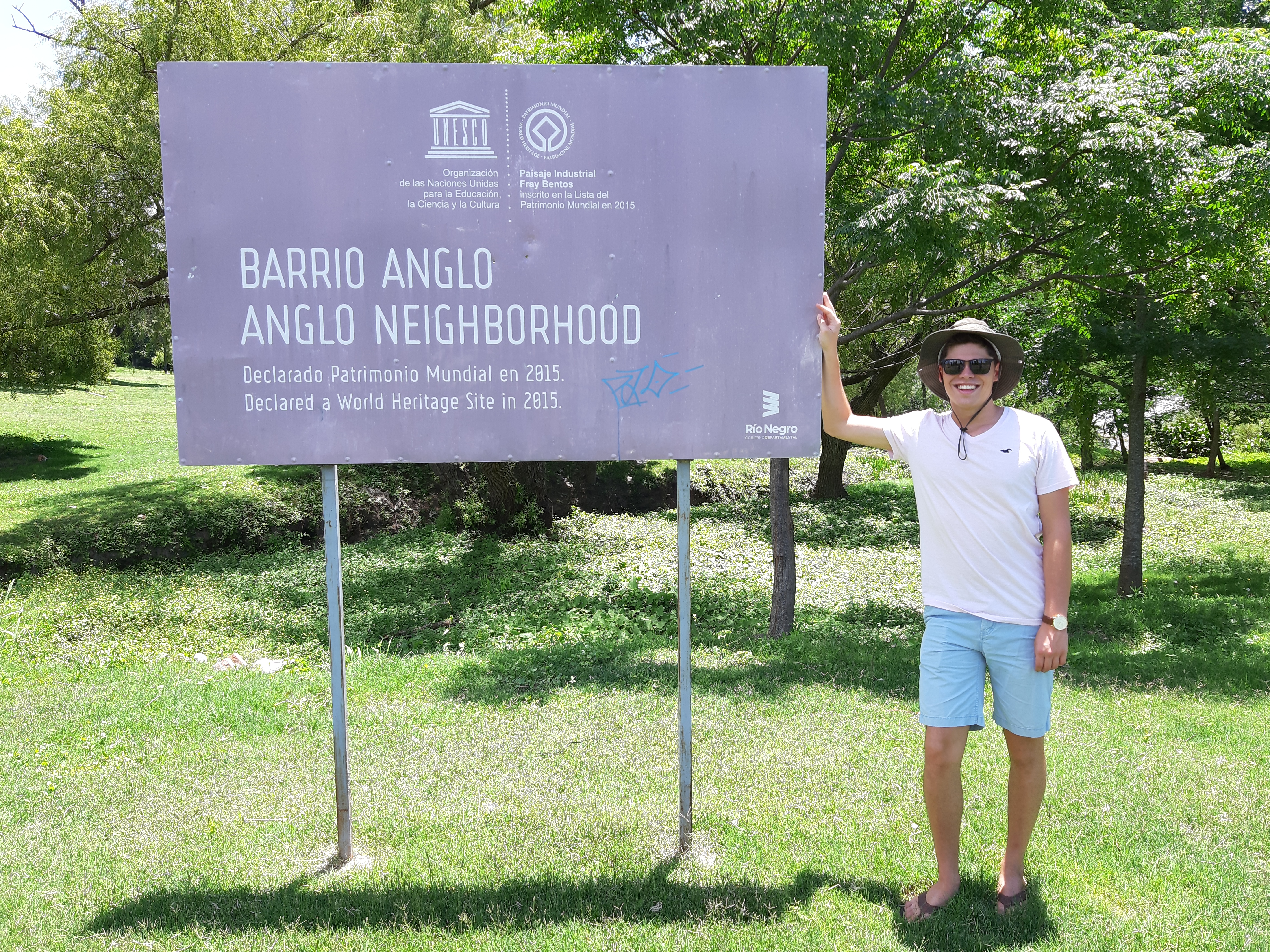 Das Bild zeigt Paulo Glowacki, der neben einem großen Schild steht, auf dem "Barrio Anglo" und "Anglo Neighborhood" steht. Das Schild informiert darüber, dass dieser Ort 2015 zum UNESCO-Weltkulturerbe erklärt wurde. Paulo trägt eine Sonnenbrille, einen Hut, ein weißes T-Shirt und blaue Shorts. Er lächelt in die Kamera und zeigt mit einer Hand auf das Schild. Die Umgebung ist grün und parkähnlich, mit vielen Bäumen im Hintergrund. Es scheint ein sonniger Tag zu sein, was die entspannte und fröhliche Stimmung