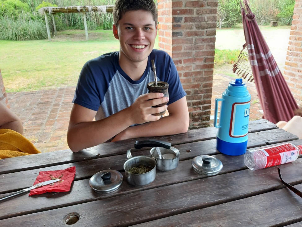 Das Bild zeigt Paulo Glowacki, der an einem Holztisch sitzt und lächelnd in die Kamera schaut. In seiner Hand hält er eine traditionelle Mate-Tasse mit einem Strohhalm, die typisch für das Trinken von Mate-Tee in Südamerika ist.