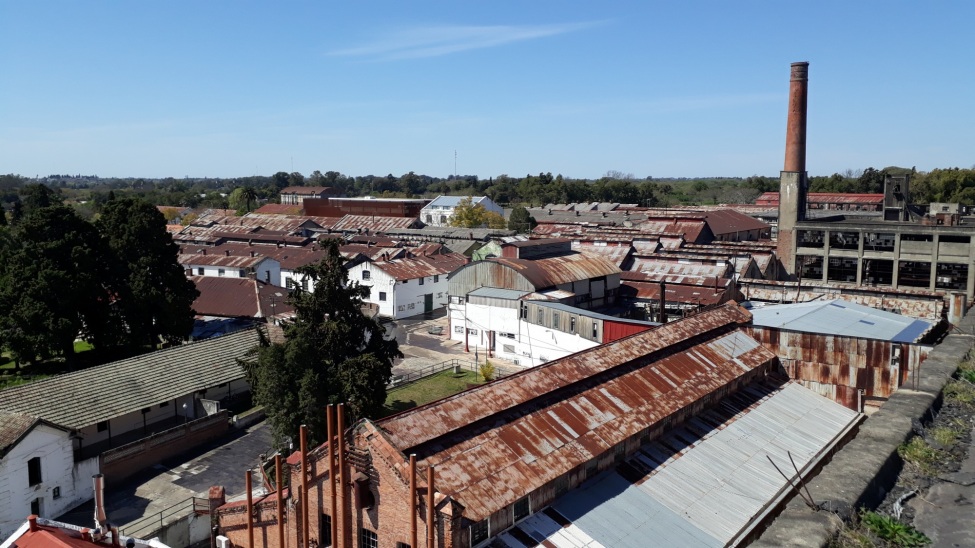 Das Bild zeigt die industrielle Landschaft von Fray Bentos mit mehreren alten Fabrikgebäuden, deren Dächer größtenteils rostig und abgenutzt sind. Im rechten Teil des Bildes ist ein hoher Schornstein zu sehen, der auf eine frühere industrielle Nutzung der Anlage hinweist. 