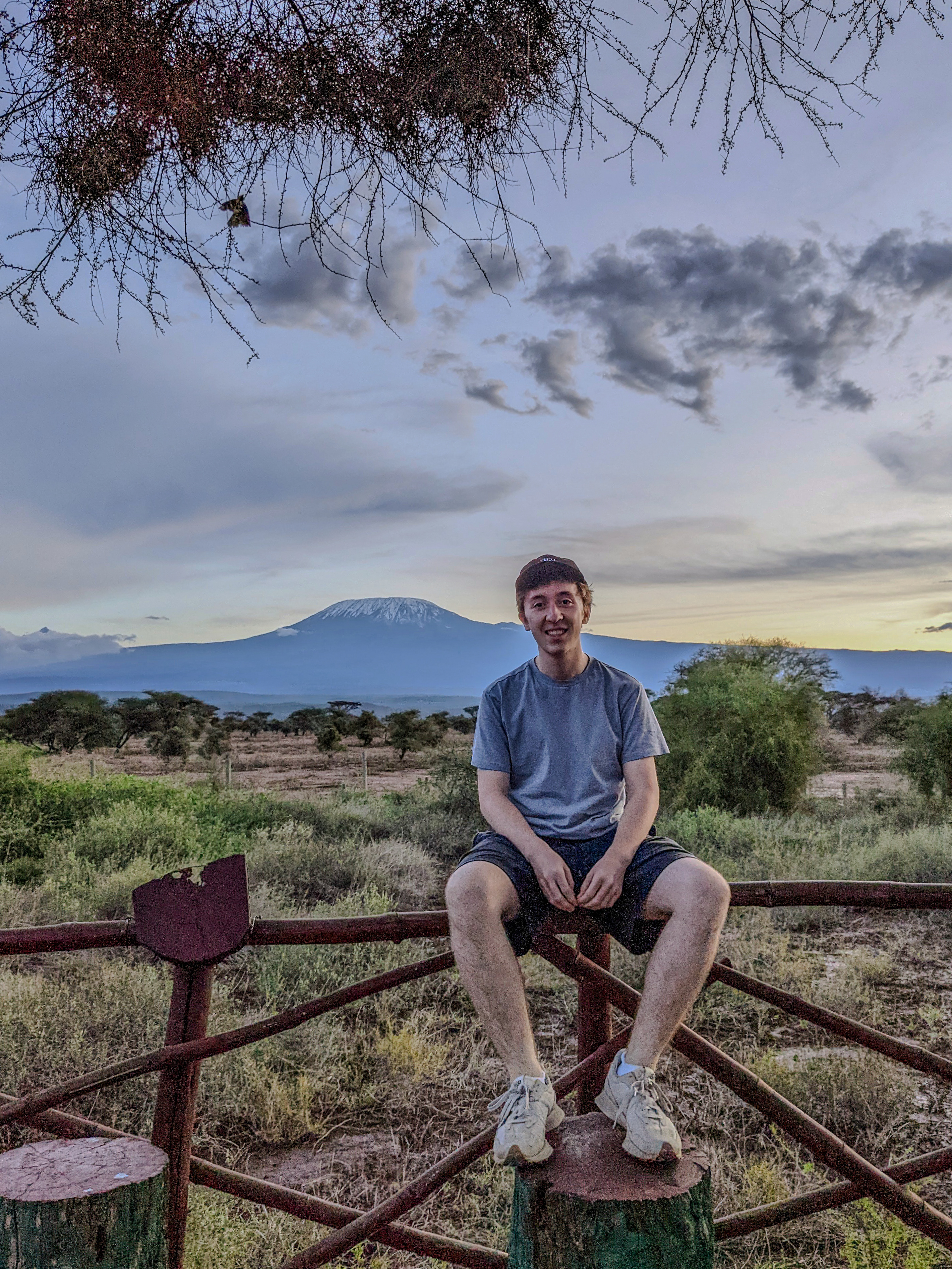 Das Bild zeigt Raphael Putz, der auf einem Holzpfosten sitzt. Im Hintergrund ist eine weite Landschaft zu sehen, die von Bäumen und Sträuchern bedeckt ist. Weiter entfernt erhebt sich ein majestätischer Berg, wahrscheinlich der Kilimandscharo, mit schneebedecktem Gipfel unter einem bewölkten Himmel. Die Atmosphäre wirkt ruhig und friedlich, und Raphael scheint die Aussicht zu genießen.