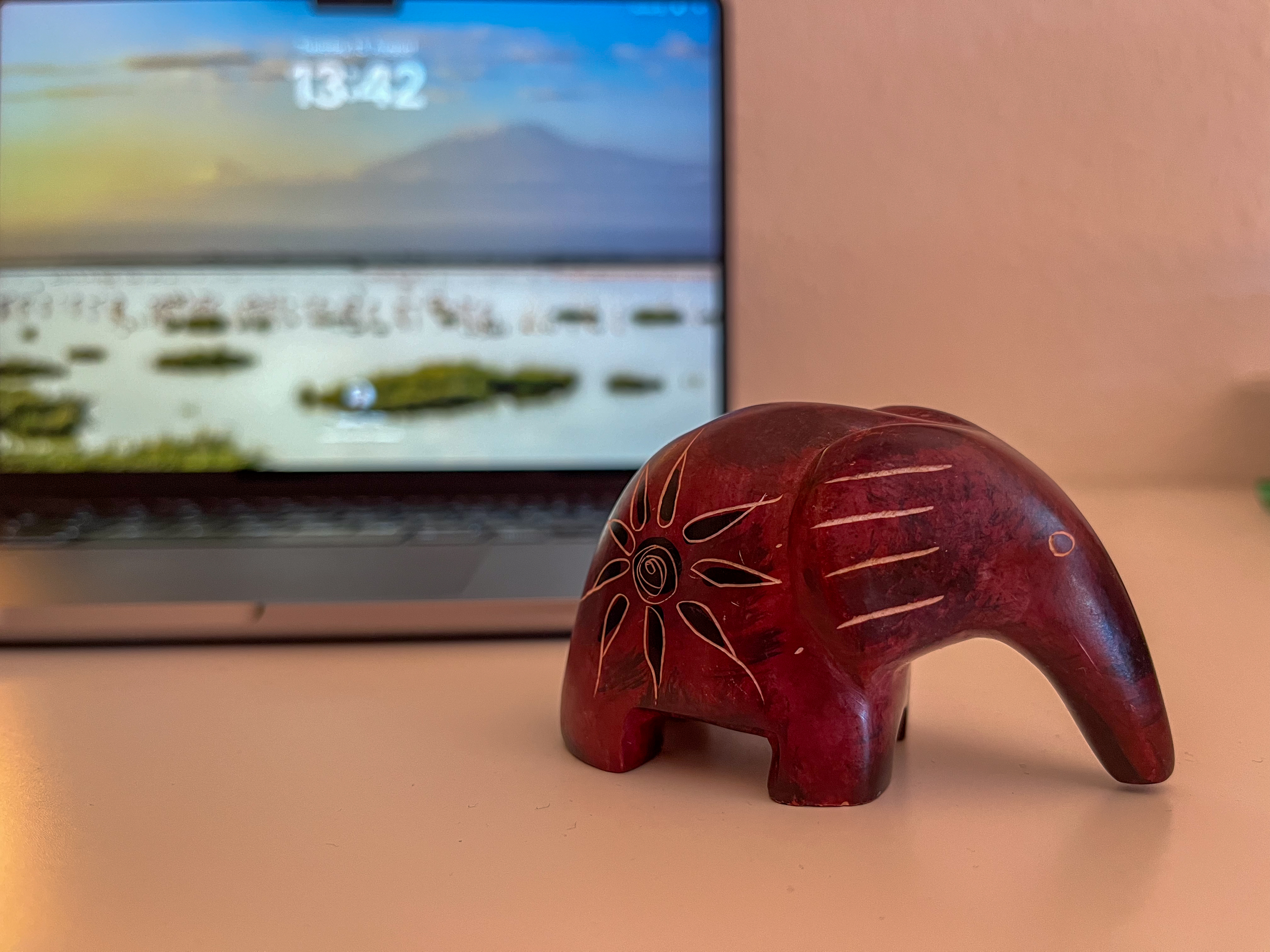 Das Bild zeigt eine kleine, rot-braune Elefantenfigur aus Holz oder Stein, die kunstvoll mit feinen Linien und Mustern verziert ist. Im Hintergrund befindet sich ein Laptop, dessen Bildschirm ein unscharfes Hintergrundbild zeigt, das vermutlich eine Landschaft mit Wasser, Vegetation und Bergen bei Sonnenuntergang darstellt. 