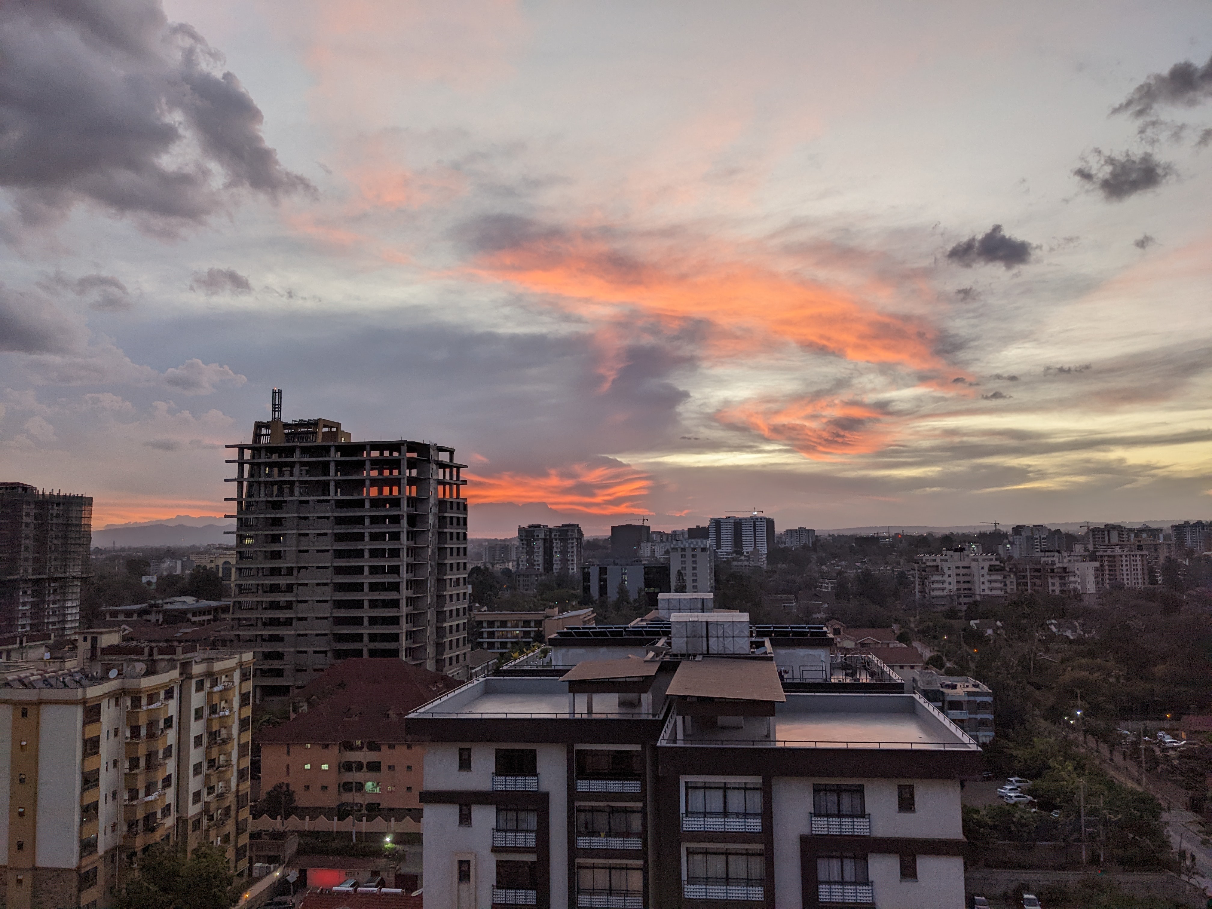 Das Bild zeigt eine städtische Skyline bei Sonnenuntergang. Im Vordergrund befinden sich mehrere Gebäude, einige im Bau, andere bereits fertiggestellt. Die Gebäude bestehen hauptsächlich aus Beton und Glas. Der Himmel ist größtenteils bedeckt, mit einem spektakulären, leuchtend orangefarbenen Wolkenband, das sich über den Horizont erstreckt und dem Himmel eine dramatische Note verleiht. Im Hintergrund sieht man weitere Gebäude, die die Stadtlandschaft prägen. 