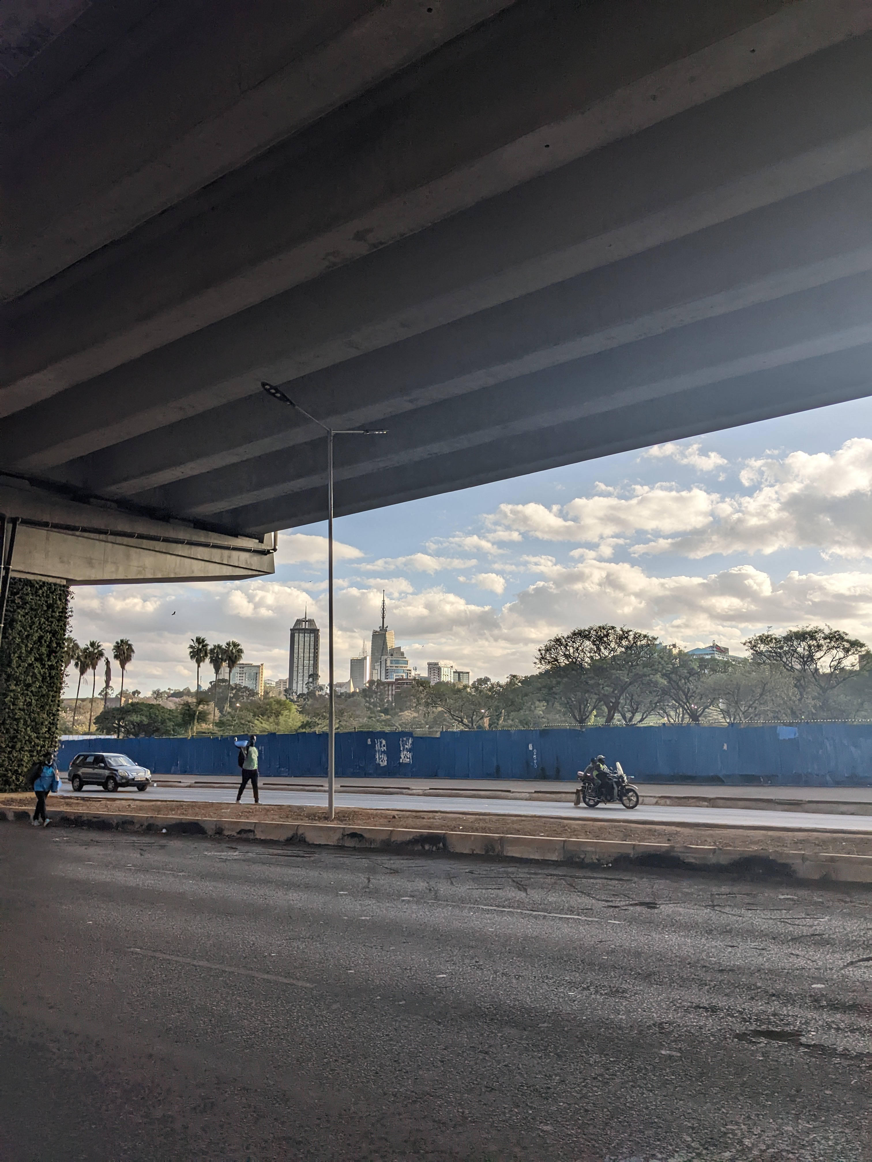 Das Bild zeigt eine Straße unter einer Brücke in einer Stadtlandschaft. Im Vordergrund verläuft die Straße, auf der ein Motorradfahrer und eine Person, die scheinbar auf den Verkehr achtet oder ihn dirigiert, zu sehen sind. Ein weiteres Fahrzeug steht am Straßenrand. Der Blick geht durch die Lücke unter der Brücke hindurch auf die Skyline der Stadt im Hintergrund. Mehrere hohe Gebäude, darunter ein Turm mit einer markanten Spitze, ragen in den Himmel, der von vereinzelten Wolken bedeckt ist. Auf der gegenüb