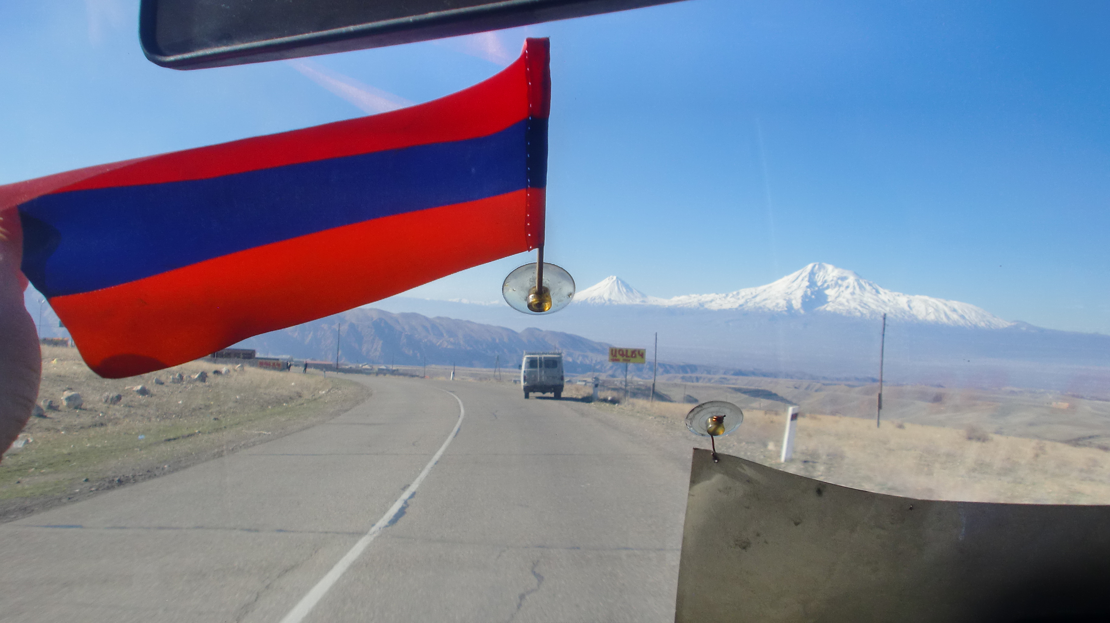 Das Bild zeigt den Blick aus einem Fahrzeug auf eine Straße, die sich durch eine karge, bergige Landschaft schlängelt. Im Vordergrund hängt eine kleine Flagge, die durch das Fahrzeugfenster befestigt ist, und im Hintergrund ragt ein schneebedeckter Berg majestätisch in den Himmel. Ein weiteres Fahrzeug ist auf der Straße voranfahrend zu sehen. Der Himmel ist klar und blau, und die Szene vermittelt ein Gefühl von Weite und Reisefreiheit. Die Flagge und die schneebedeckten Gipfel im Hintergrund könnten darauf