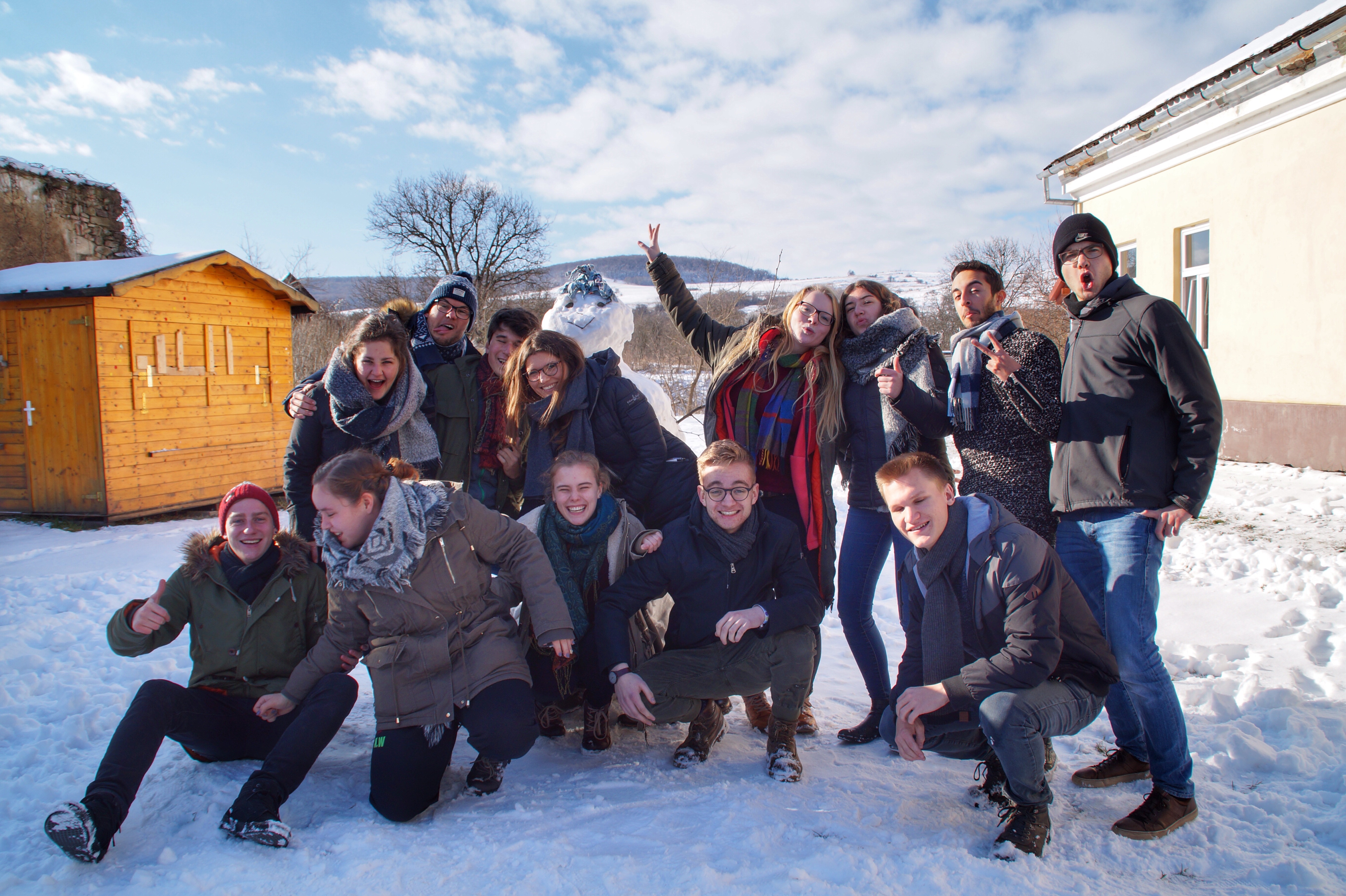 Das Bild zeigt eine fröhliche Gruppe von jungen Menschen, die im Schnee vor einer winterlichen Landschaft posieren. Die Gruppe ist in warme Winterkleidung gehüllt, darunter dicke Jacken, Schals und Mützen. Einige Personen lachen, während andere lustige Posen einnehmen oder Zeichen mit den Händen machen. Im Hintergrund ist ein kleines Holzhaus zu sehen, und die Umgebung wirkt ländlich, mit schneebedeckten Hügeln und Bäumen. 