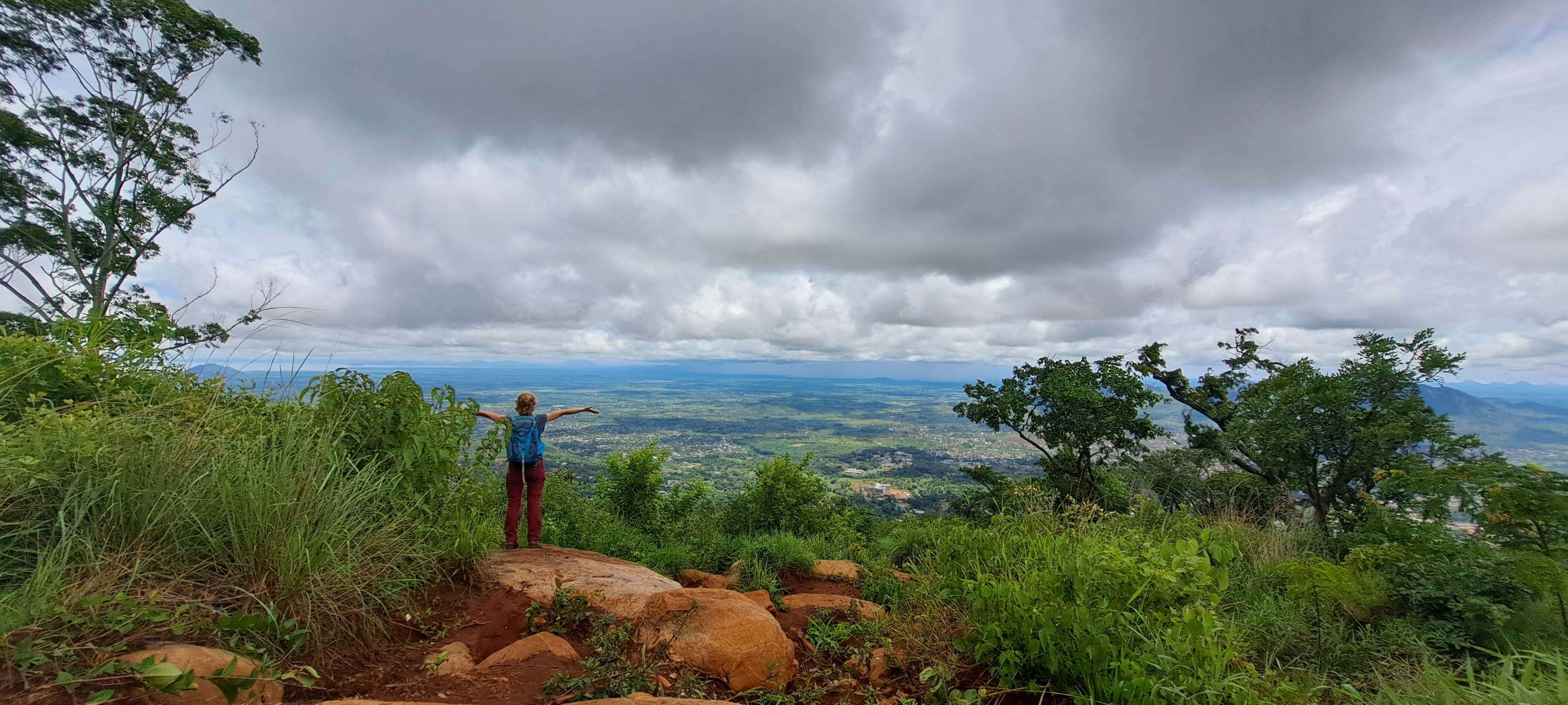 Das Bild zeigt eine Person mit einem blauen Rucksack, die auf einem felsigen Vorsprung steht und die Arme weit ausbreitet, als würde sie die Aussicht auf die darunter liegende, weitläufige Landschaft begrüßen. Die Szene ist in einer grünen, üppigen Umgebung mit Bäumen und Sträuchern aufgenommen. Der Himmel ist größtenteils bewölkt, was der Landschaft eine dramatische und zugleich friedliche Atmosphäre verleiht. Im Hintergrund erstreckt sich ein weites Tal mit Feldern und möglicherweise einer Stadt in der Fe