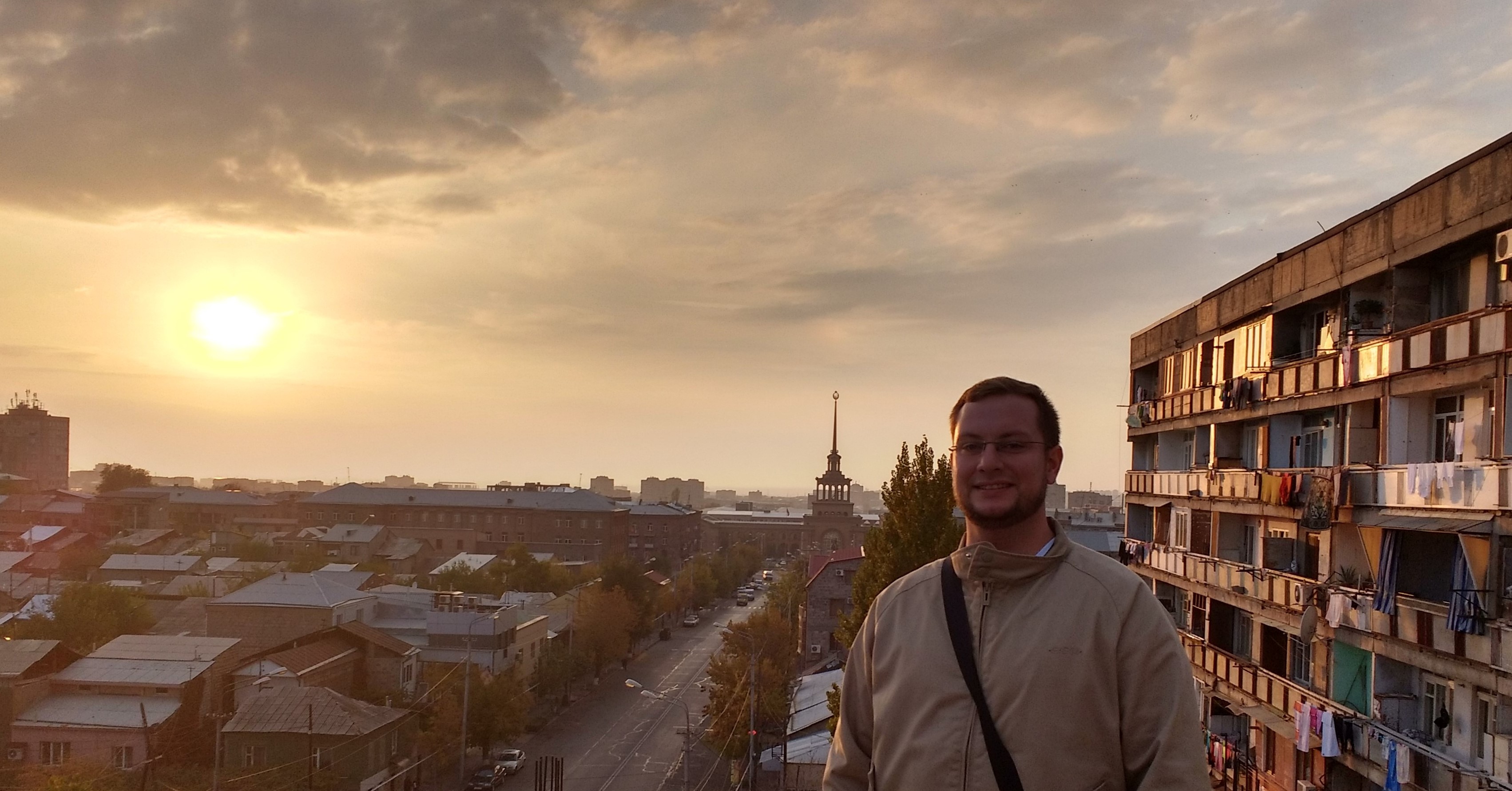 Das Bild zeigt einen Mann, der auf einer erhöhten Position in einer Stadt steht, während die Sonne im Hintergrund untergeht und die Szene in warmes, goldenes Licht taucht. Der Mann trägt eine beige Jacke und lächelt in die Kamera. Im Hintergrund erstreckt sich eine Stadtlandschaft mit verschiedenen Gebäuden, darunter ein markantes Gebäude mit einem Turm und einer Spitze. Auf der rechten Seite des Bildes ist ein mehrstöckiges Wohngebäude zu sehen, an dem Wäsche auf den Balkonen hängt. Die Stimmung des Bildes