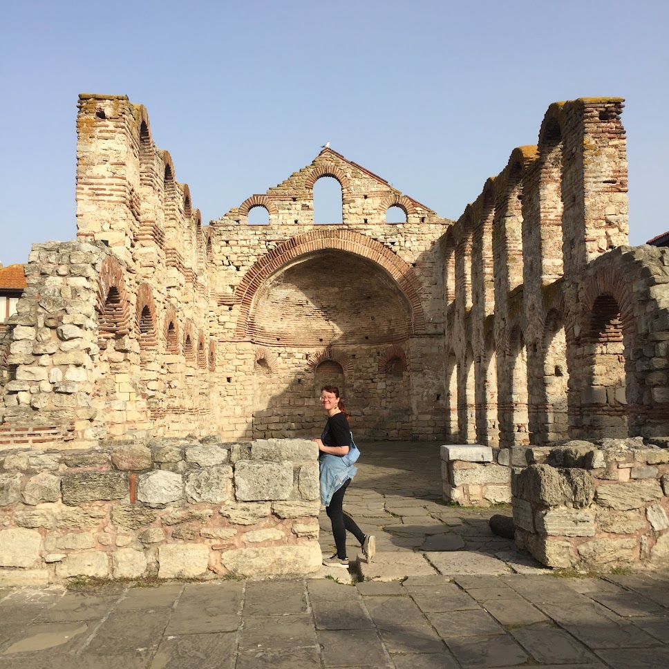 Das Bild zeigt eine Frau, die durch die Ruinen einer alten Kirche oder eines historischen Gebäudes geht. Die Struktur besteht aus alten Steinmauern mit Bögen und Fensteröffnungen, die teilweise erhalten geblieben sind. Die Frau trägt eine schwarze Oberbekleidung und hat eine Jacke um die Taille gebunden. Sie blickt über ihre Schulter zurück zur Kamera und lächelt. Der Boden besteht aus großen Steinplatten, und die Szenerie ist bei Tageslicht aufgenommen, was die Details der Ruinen gut sichtbar macht. 