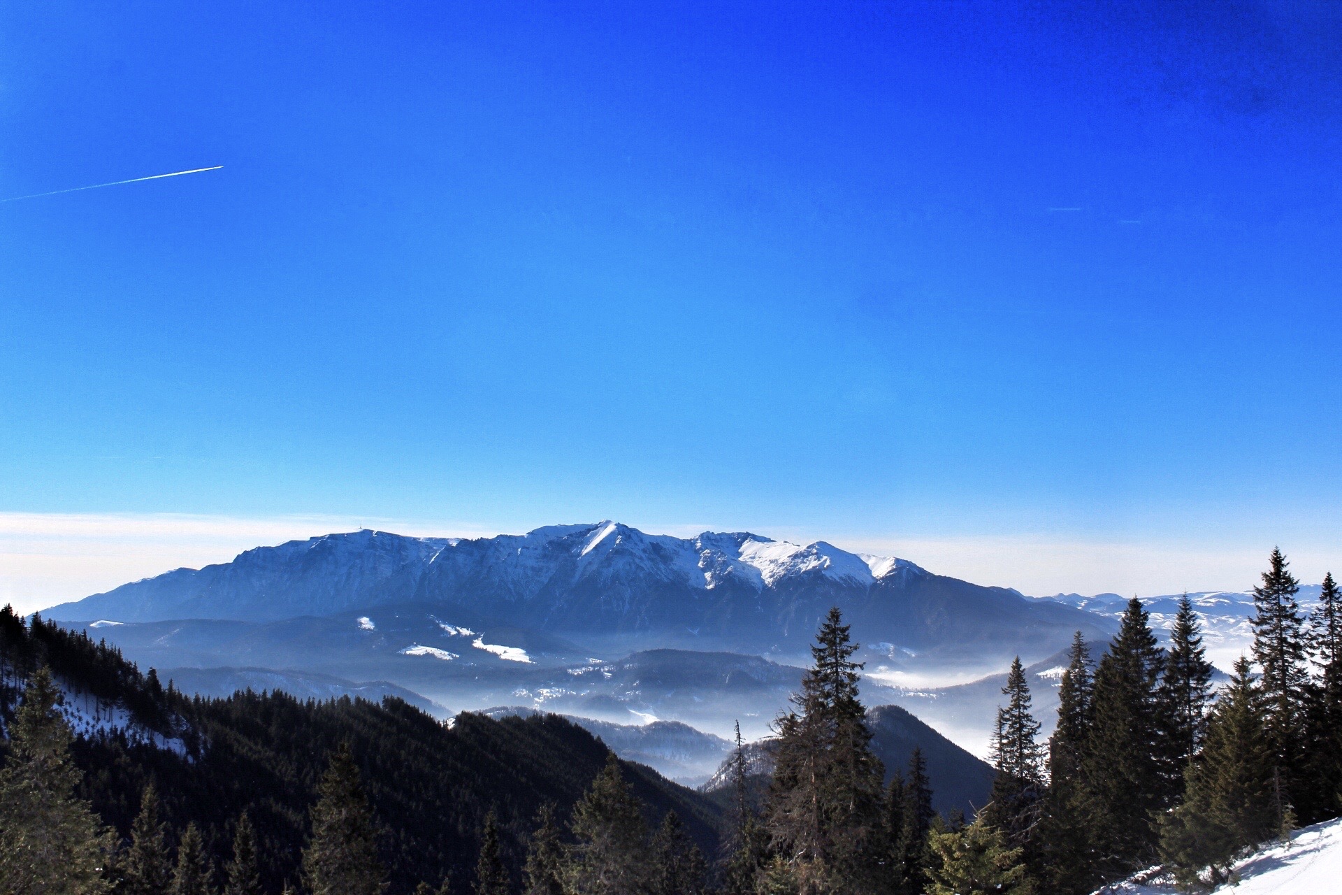 Das Bild zeigt eine beeindruckende Berglandschaft mit schneebedeckten Gipfeln unter einem strahlend blauen Himmel. Im Vordergrund sind dunkle, bewaldete Hügel zu sehen, die in das Tal hinabführen, während die schneebedeckten Berge im Hintergrund majestätisch aufragen. Die klaren Linien der Berge heben sich scharf gegen den Himmel ab, und die gesamte Szenerie strahlt eine ruhige, kühle Atmosphäre aus, die typisch für Winterlandschaften in den Bergen ist. Ein Kondensstreifen eines Flugzeugs ist am linken ober