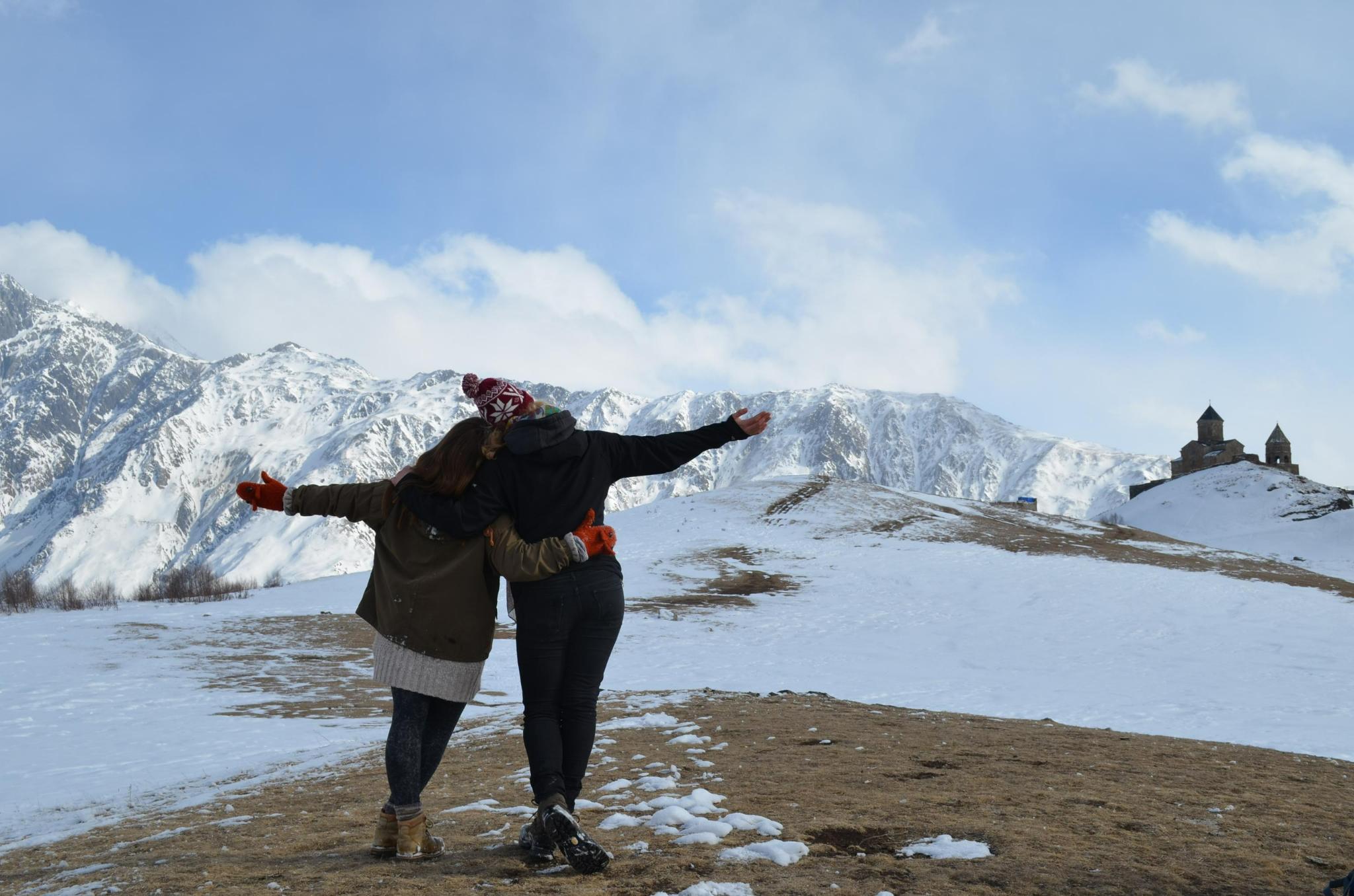 Das Bild zeigt zwei Personen von hinten, die sich umarmen und gemeinsam auf eine schneebedeckte Berglandschaft blicken. Sie stehen auf einem Hügel, der teilweise mit Schnee bedeckt ist, und strecken ihre Arme in einer offenen, freudigen Geste aus. Die beiden tragen warme Kleidung, darunter Mützen und Handschuhe, die für die winterlichen Bedingungen geeignet sind. Im Hintergrund ist eine alte, kleine Kirche oder Kapelle zu sehen, die auf einem weiteren Hügel steht und sich deutlich vor den imposanten, schnee