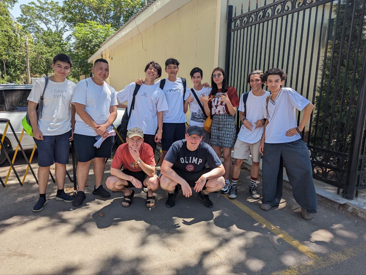 Das Bild zeigt eine Gruppe von jungen Menschen, die in einer lockeren Pose zusammenstehen und lächeln. Einige der Personen tragen weiße T-Shirts und Shorts, während andere lässige Sommerkleidung anhaben. Zwei Personen hocken in der vorderen Reihe, während die anderen hinter ihnen stehen. Im Hintergrund ist ein großes Metalltor zu sehen, das zu einem Gebäude führt, möglicherweise eine Schule oder ein Gemeindezentrum. 