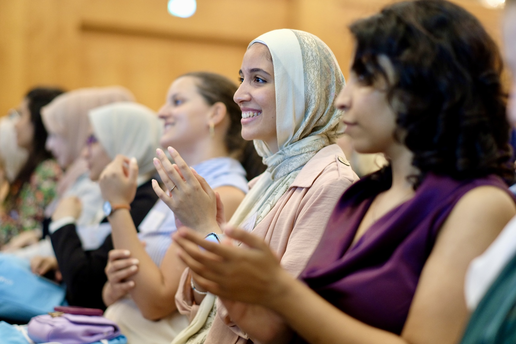 Eine Gruppe von Frauen sitzt in einem Veranstaltungsraum und applaudiert lächelnd. Eine Frau mit hellem Kopftuch in der Mitte des Bildes strahlt, während sie klatscht.