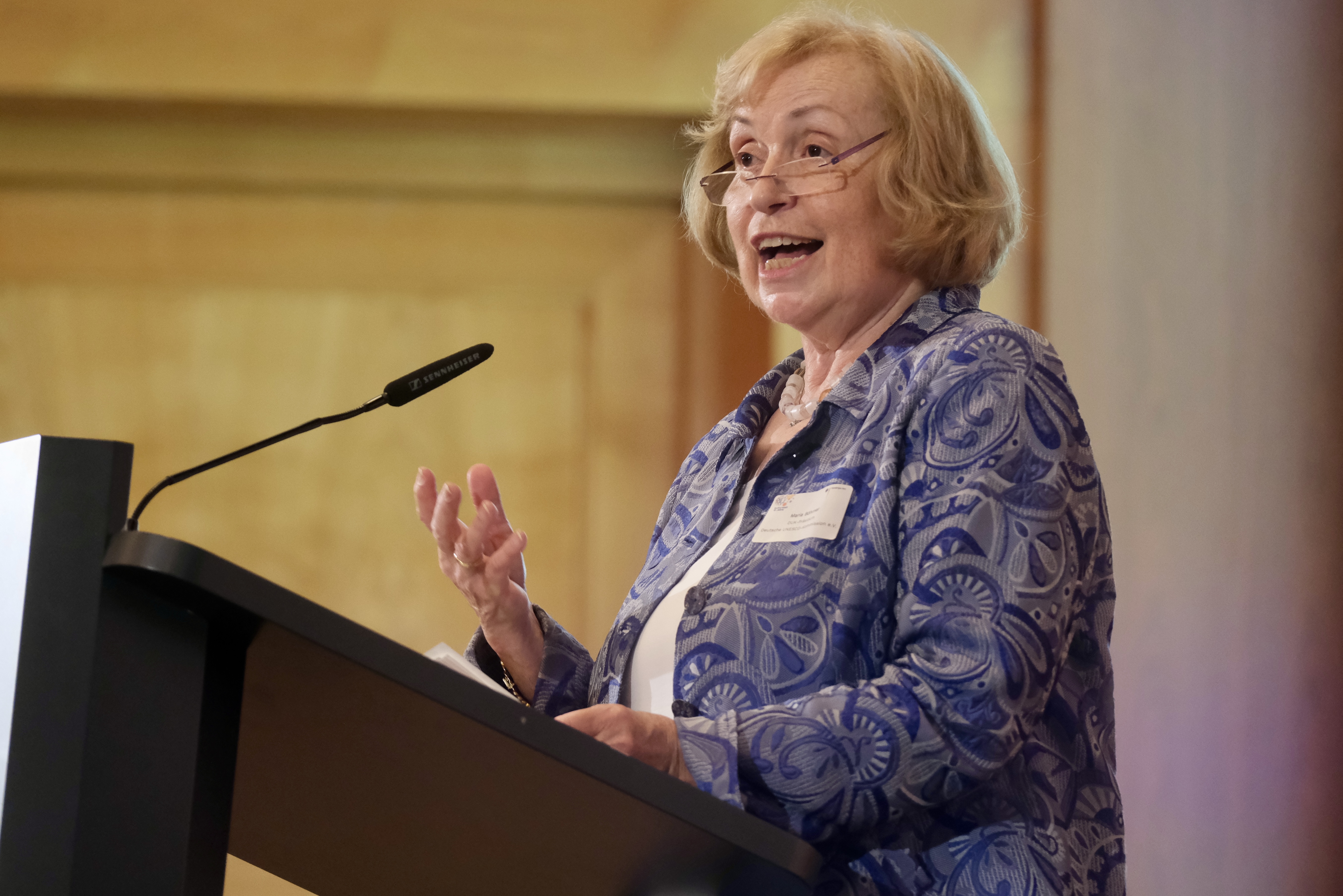 Die Präsidentin der Deutschen UNESCO Kommission, Maria Böhmer, steht vor dem Podest und hält eine Rede. Sie hat blondes kurzes Haar, trägt eine Brille und eine blaue Bluse.