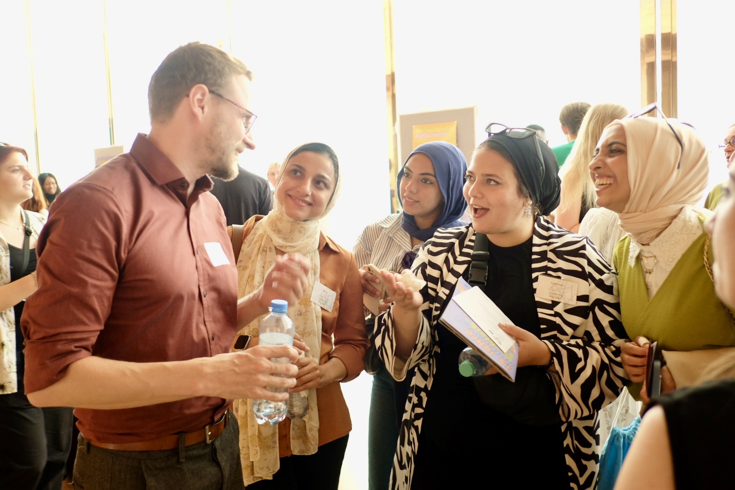 Eine Gruppe von fünf Personen, darunter ein Mann und vier Frauen mit Kopftuch, unterhalten sich angeregt bei einem Empfang. Der Mann trägt ein braunes Hemd und hält eine Wasserflasche, während die Frauen aufmerksam zuhören und lächeln.