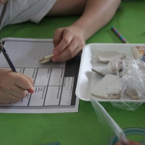 Das Foto zeigt eine Kinderhand, die in der linken Hand eine Tonscherbe hält und mit der rechten eine Notiz auf einem Papier verfasst.