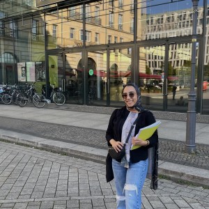 Farah steht lächelnd mit Sonnenbrille vor der Glasfassade der Akademie der Künste