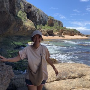 Anne steht in einem hellen T-Shirt und einer Kappe auf felsigem Gelände an einem Küstestreifen. Sie hält sich an einem Felsen fest, während im Hintergrund eine malerische Klippenlandschaft und das Meer zu sehen sind. Der Himmel ist klar und blau.