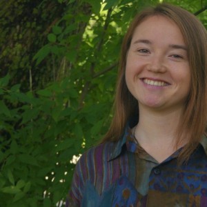 Das Bild zeigt Mara Urich. Sie hat lange, glatte, braune Haare und lächelt in die Kamera. Sie trägt ein bunt gemustertes Hemd. Im Hintergrund sind grüne Blätter und ein Baumstamm zu sehen, was auf eine Wald- oder Gartenumgebung hinweist.