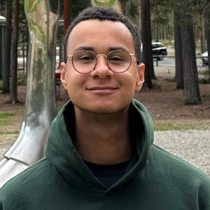 Philipp Mensah lächelt leicht in die Kamera. Er hat kurze schwarze Hare und trägt eine Brille sowie einen grünen Kapuzenpullover. Der Hintergrund zeigt einen Wald mit hohen Bäumen und einem kunstvollen, silbernen Objekt, sowie einige geparkte Autos in der Ferne. Der Boden besteht aus Schotter und Grasflächen.