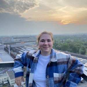 Das Bild zeigt Pauline Rothkegel, die auf einem Balkon oder einer erhöhten Plattform steht. Sie trägt eine karierte, flauschige Jacke in Blautönen und ein weißes T-Shirt. Pauline hat blonde Haare, die zu einem niedrigen Zopf gebunden sind, und sie lächelt leicht in die Kamera. Im Hintergrund ist eine weite Landschaft zu sehen, mit einem Industriegebiet oder einer Fabrik im Vordergrund und Bäumen sowie einem leicht bewölkten Himmel bei Sonnenuntergang im Hintergrund.