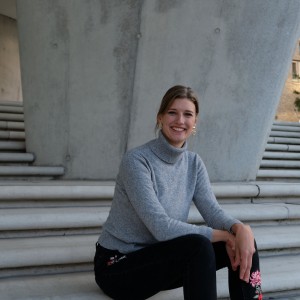 Das Bild zeigt die Alumna Bettina Gschwend. Sie trägt ihre Haare zusammengebunden und einen grauen Rollkragenpullover. Sie sitzt auf einer Betontreppe.